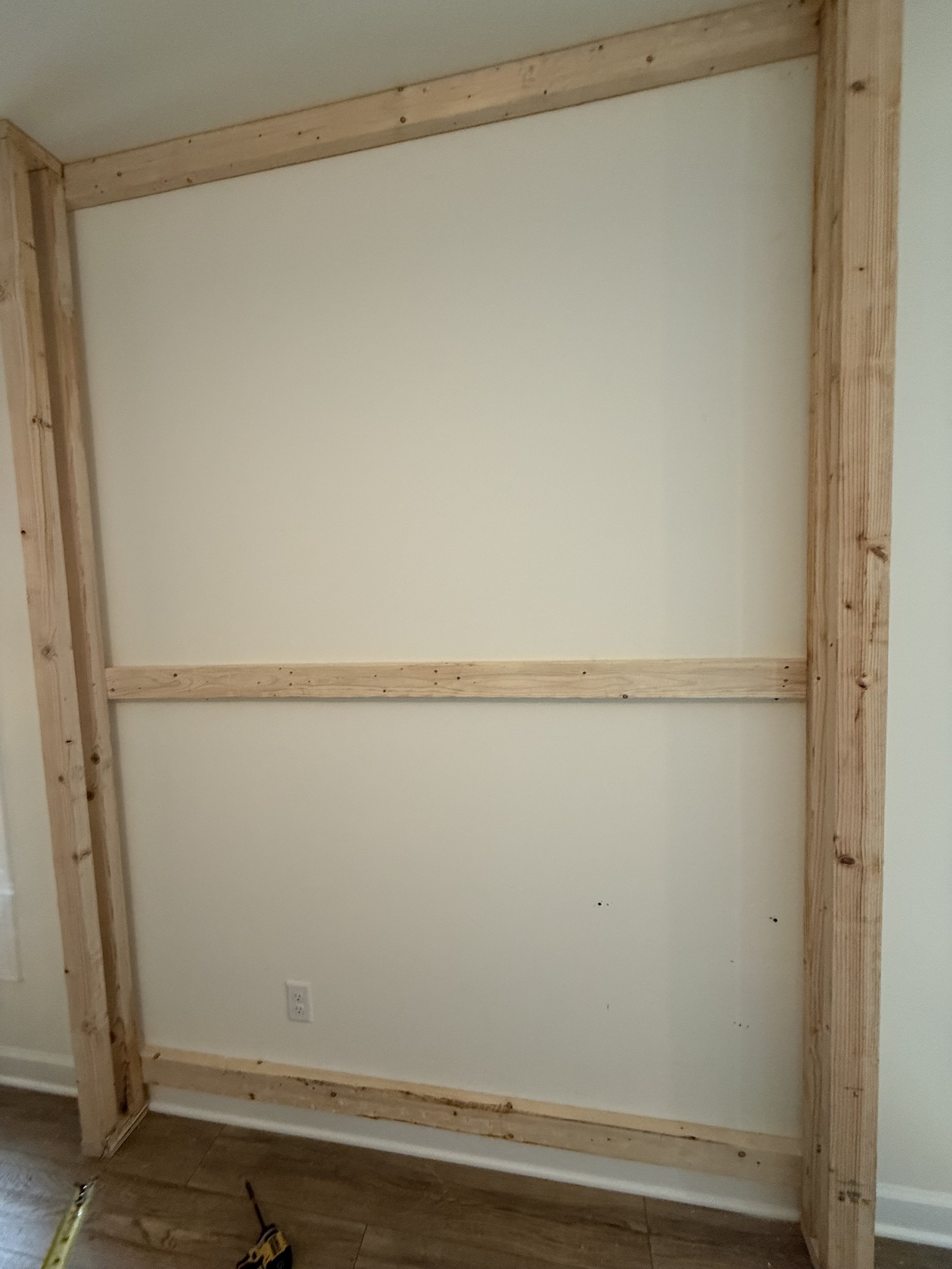 Framed wall with wooden studs and horizontal support beams, located in a room with light-colored walls and hardwood flooring.