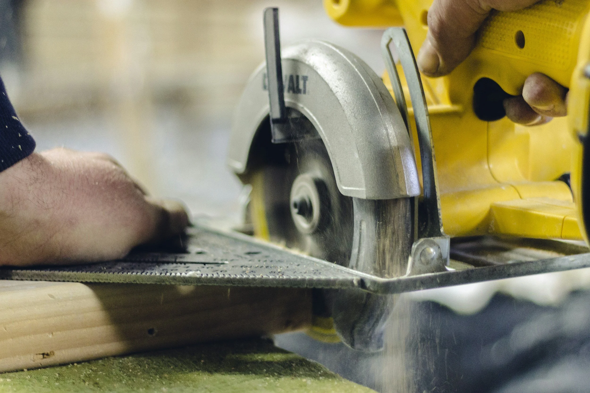 Close-up of a person using a yellow circular saw to cut a piece of wood, with the person's hands holding the wood steady.