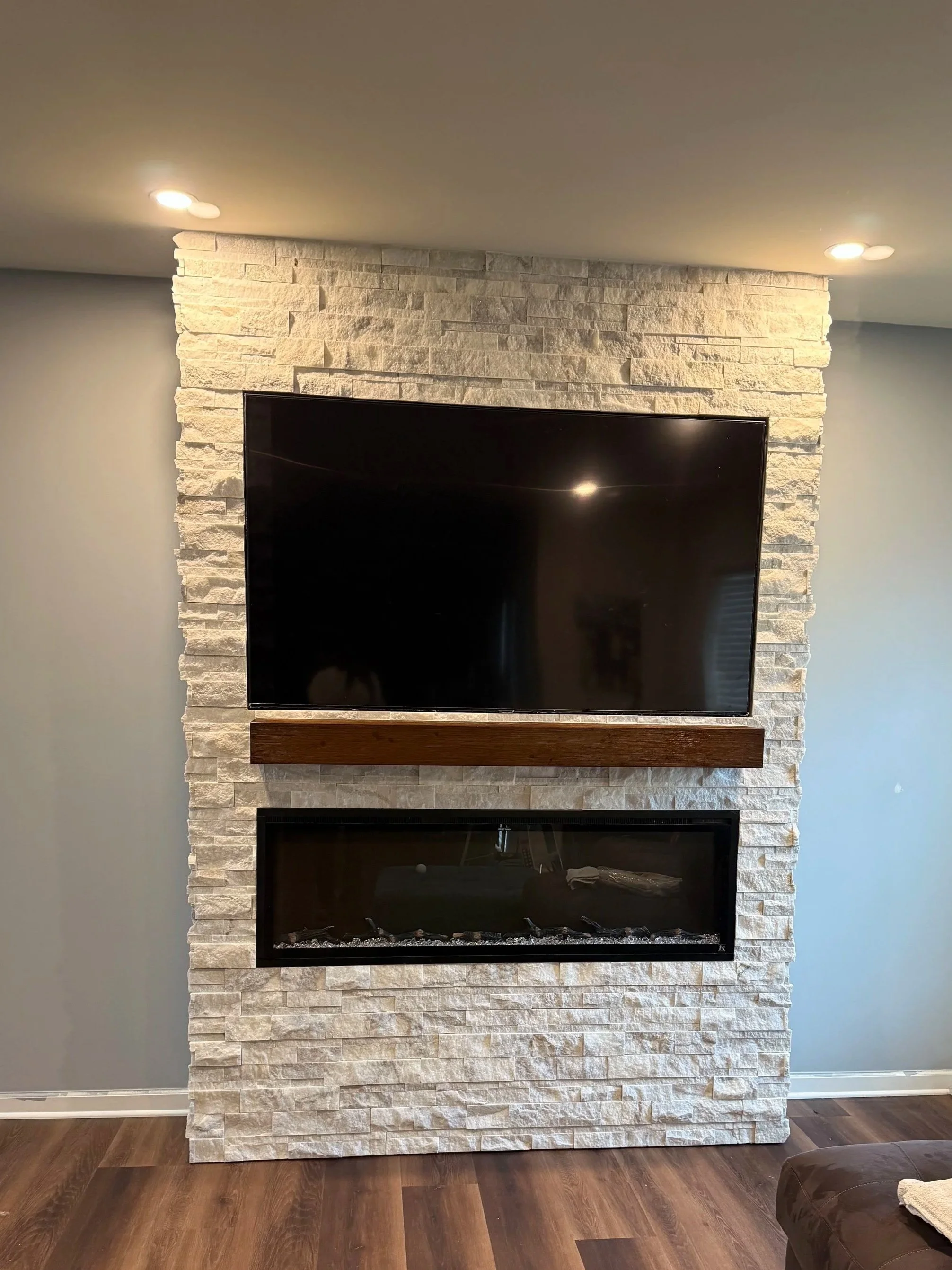 Living room with a stone fireplace, mounted flat-screen TV above a wooden mantel, and hardwood flooring. Part of a dark sofa with a towel and some items on it is visible on the right.