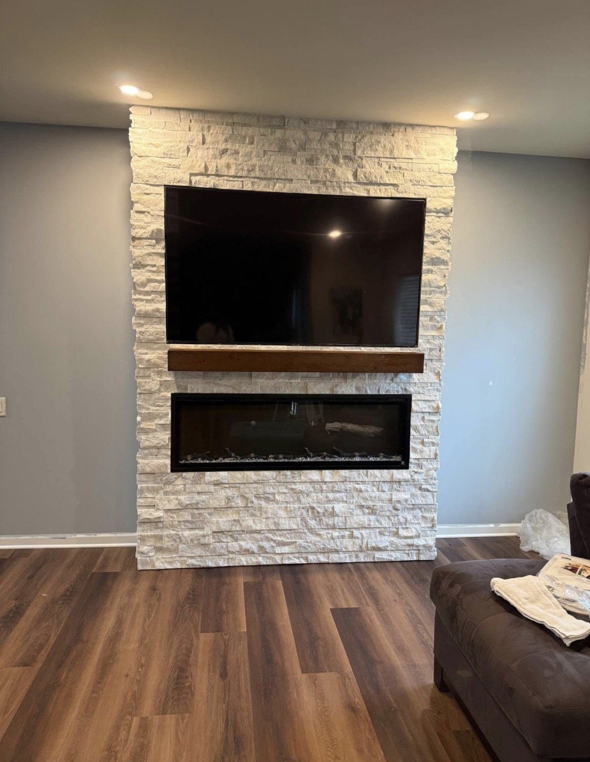Living room with a stone fireplace, mounted TV, and wooden mantel. Hardwood flooring and a brown sofa with a white towel on it.