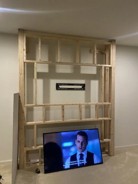 Wooden frame structure in a room with a mounted television displaying a man in a suit with subtitles.