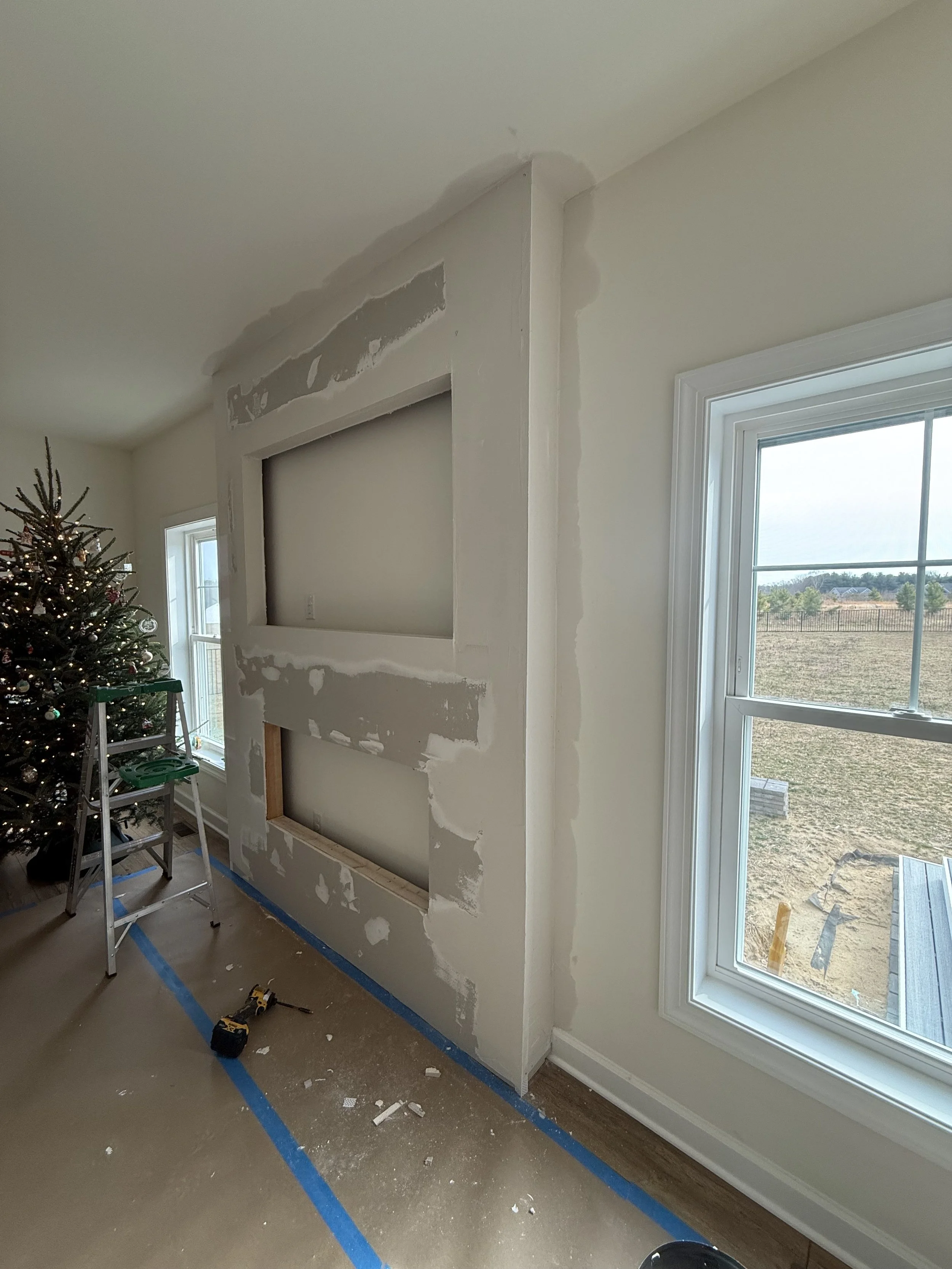 Interior wall under construction with drywall patching and framing, next to a window and decorated Christmas tree.