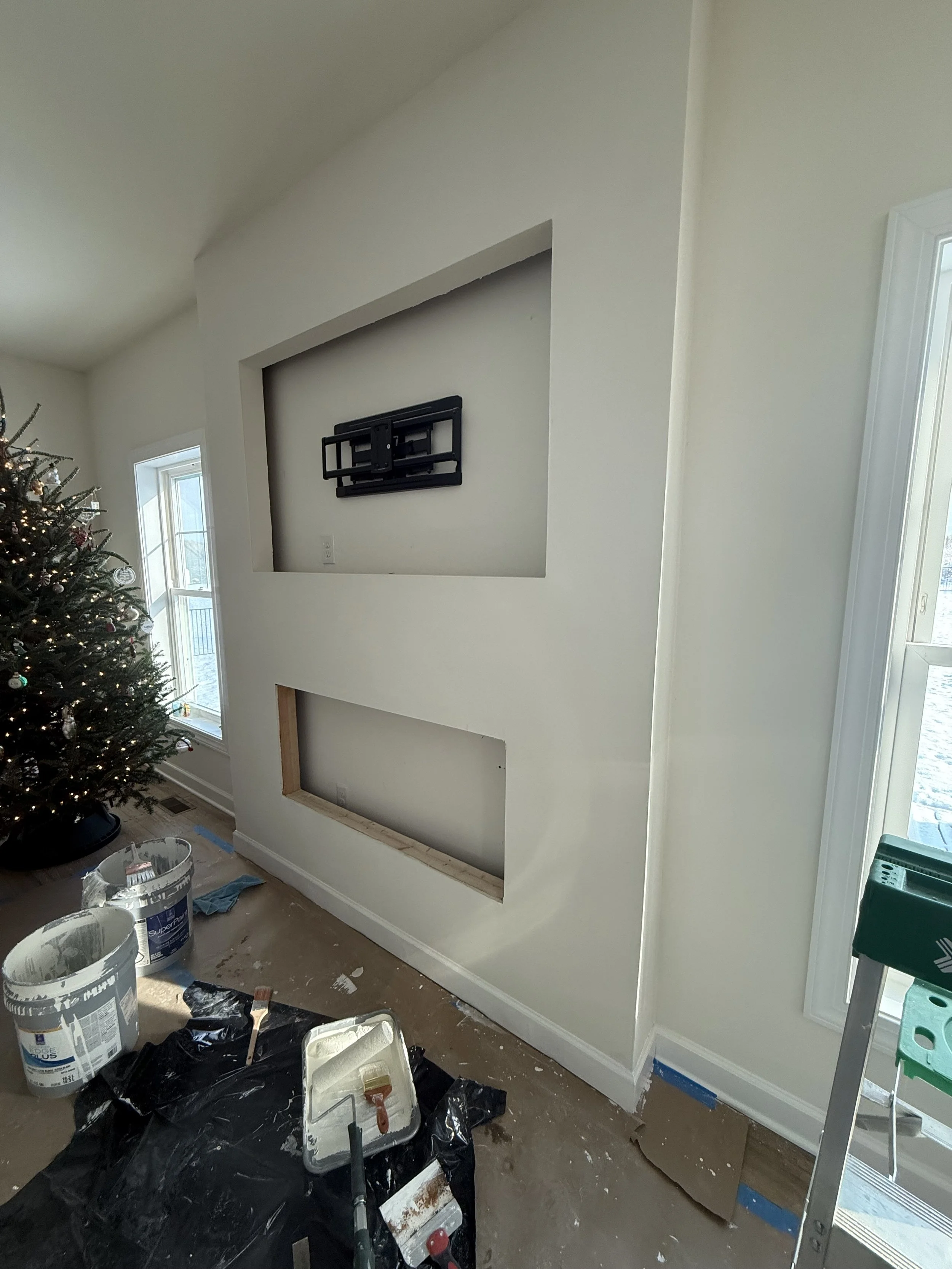Living room under renovation with an unfinished fireplace, a decorated Christmas tree, and painting supplies on the floor.