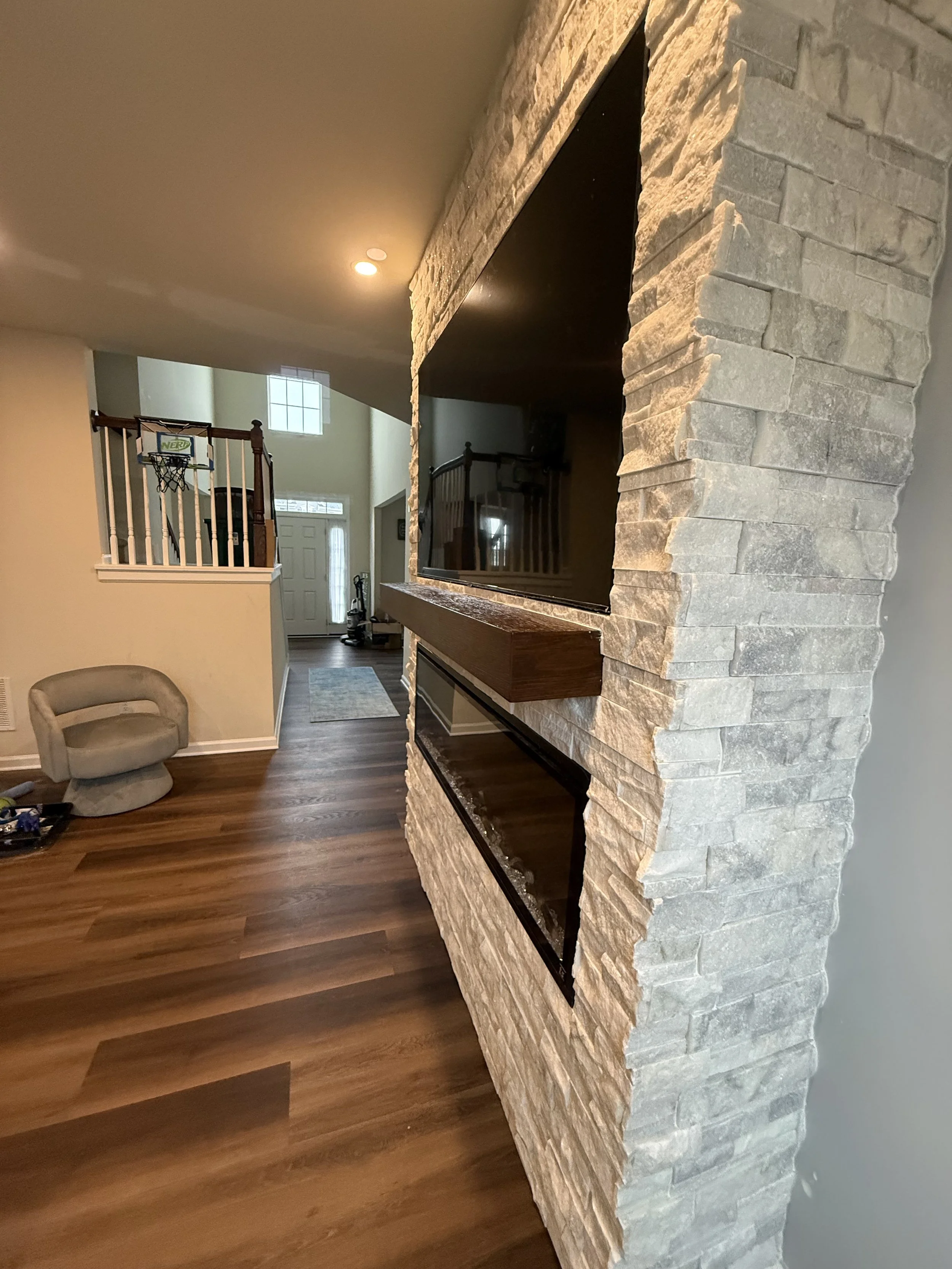 Interior of a living room showing a stone fireplace, a beige armchair, a hardwood floor, and a staircase with a basketball hoop in the background.