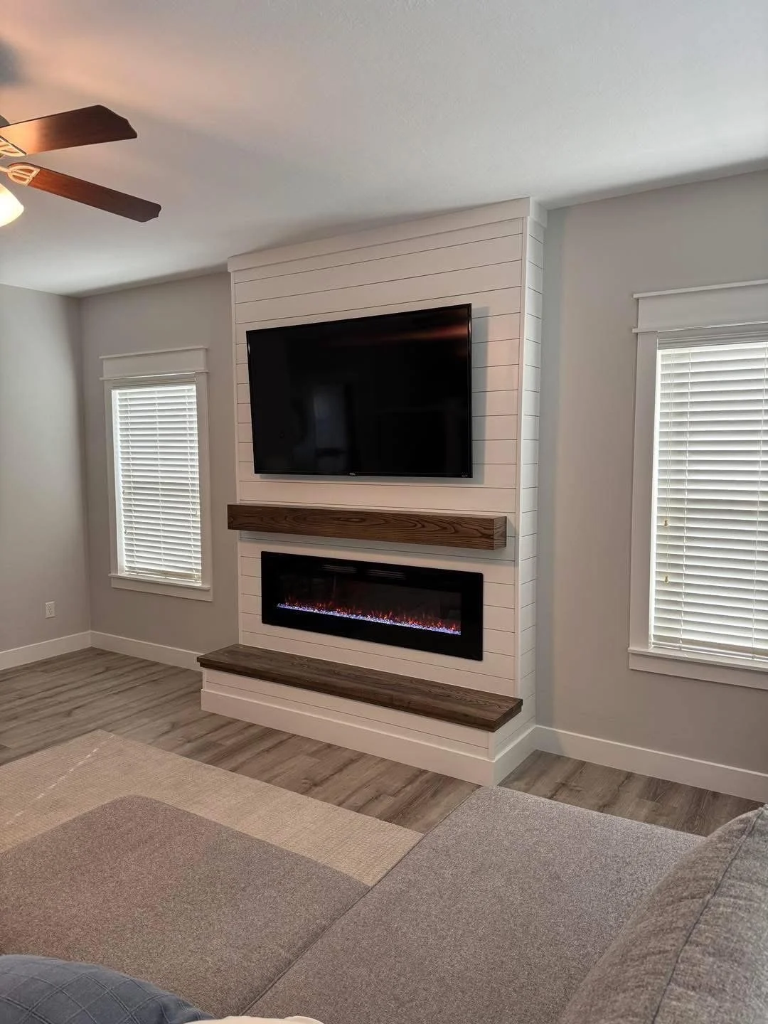 Living room with a wall-mounted TV above an electric fireplace with a wooden mantel, two windows with blinds, ceiling fan, beige and gray furniture, and light wood flooring.