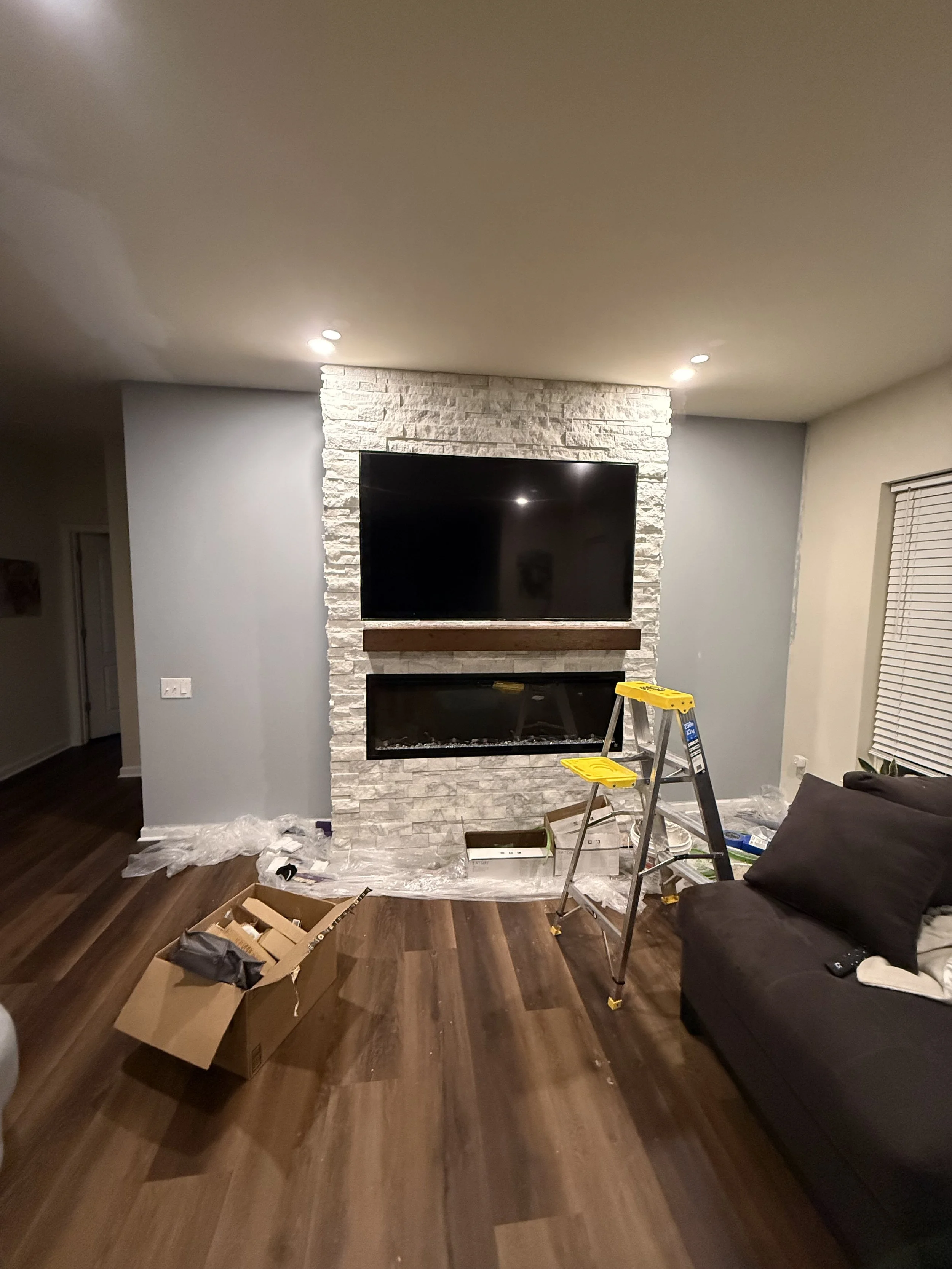 Living room interior with a stone fireplace, mounted flat-screen TV, a gray sofa, a small yellow step ladder, and construction materials scattered on the floor.