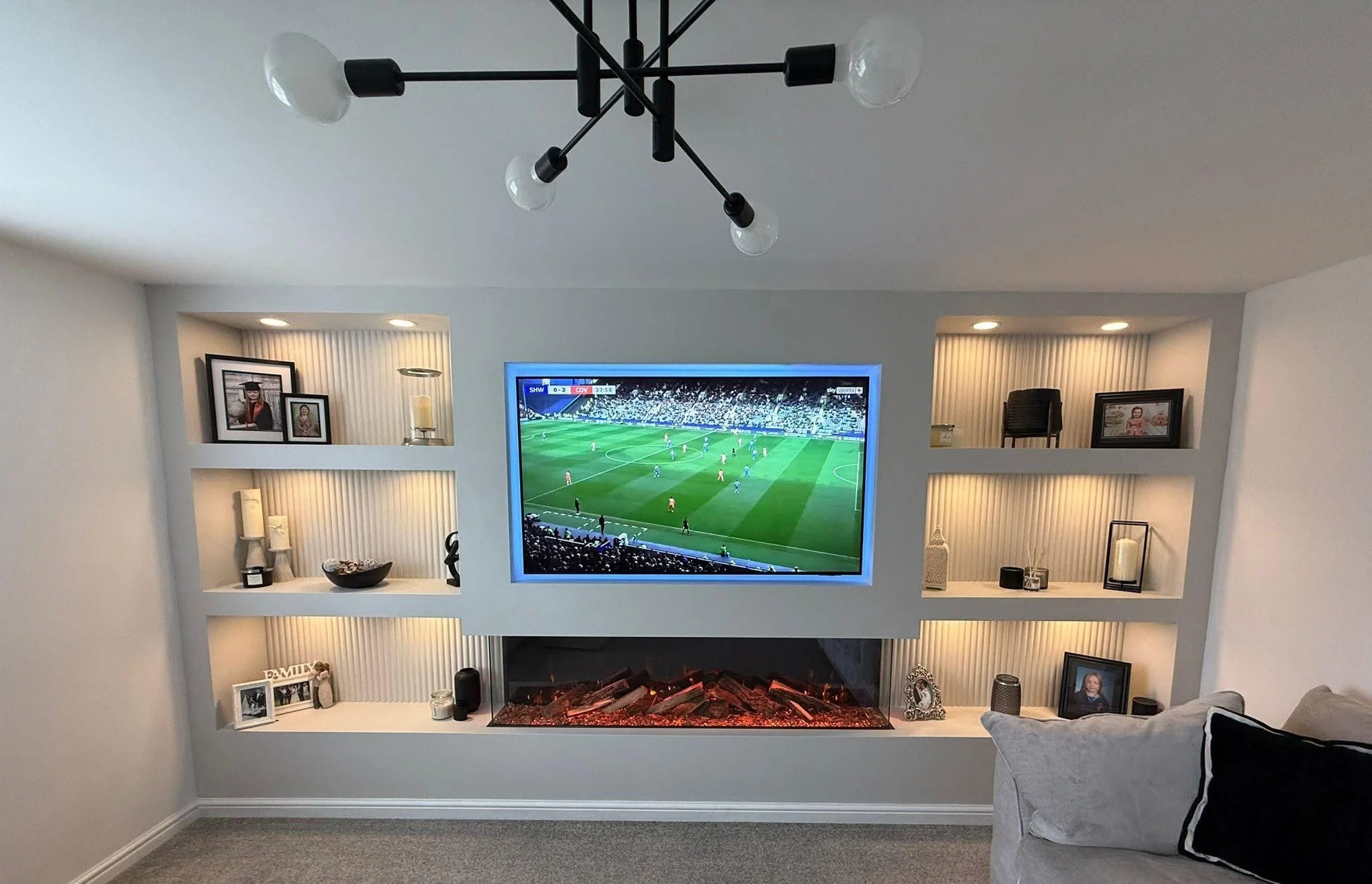 Living room with television displaying a soccer match, built-in shelves with decorative items and picture frames, electric fireplace, modern ceiling light fixture, and a beige couch with pillows.