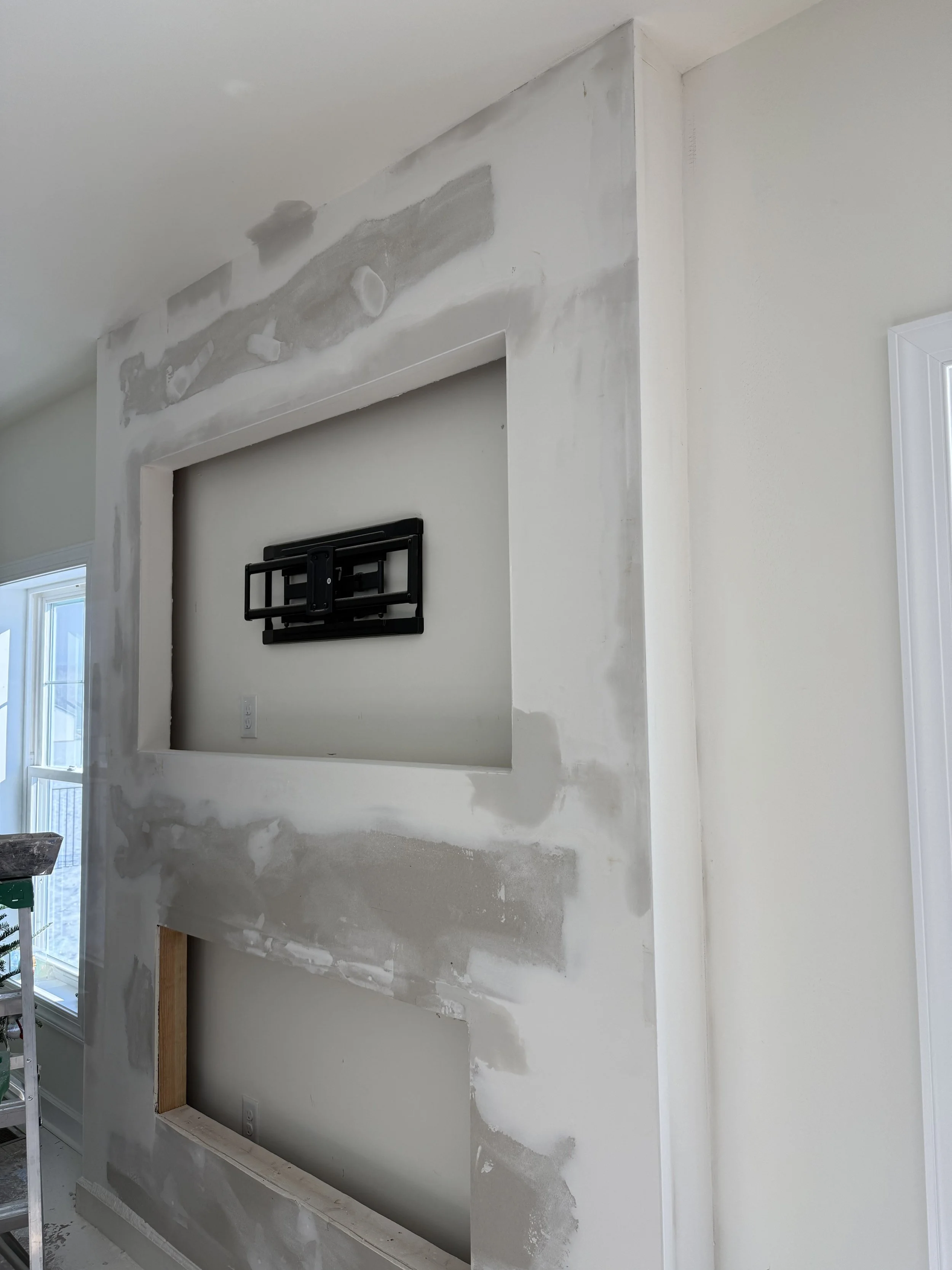 Partial drywall installation and patching on a wall surrounding a recessed area with a TV mount, indicating ongoing home renovation or construction.
