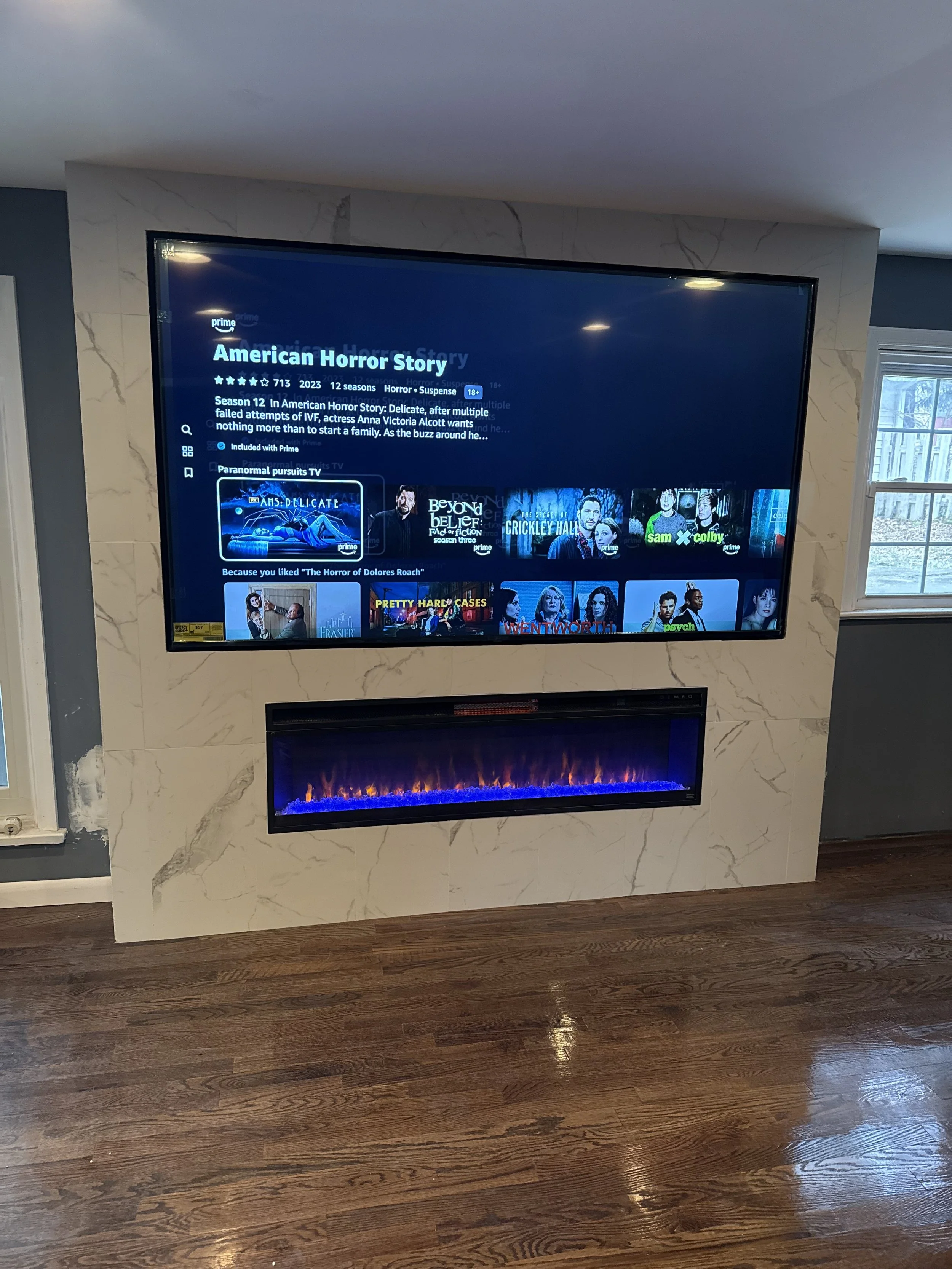 A wall-mounted television displaying the Netflix page for American Horror Story, with a fireplace below it on a marble accent wall.