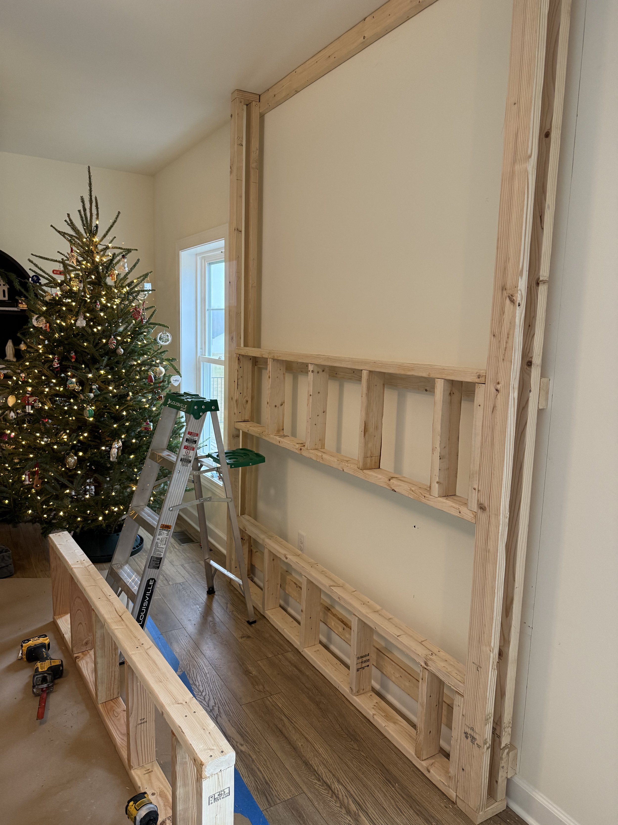 Interior of a home with a Christmas tree and a wooden wall frame under construction, with a stepladder and tools on the floor.