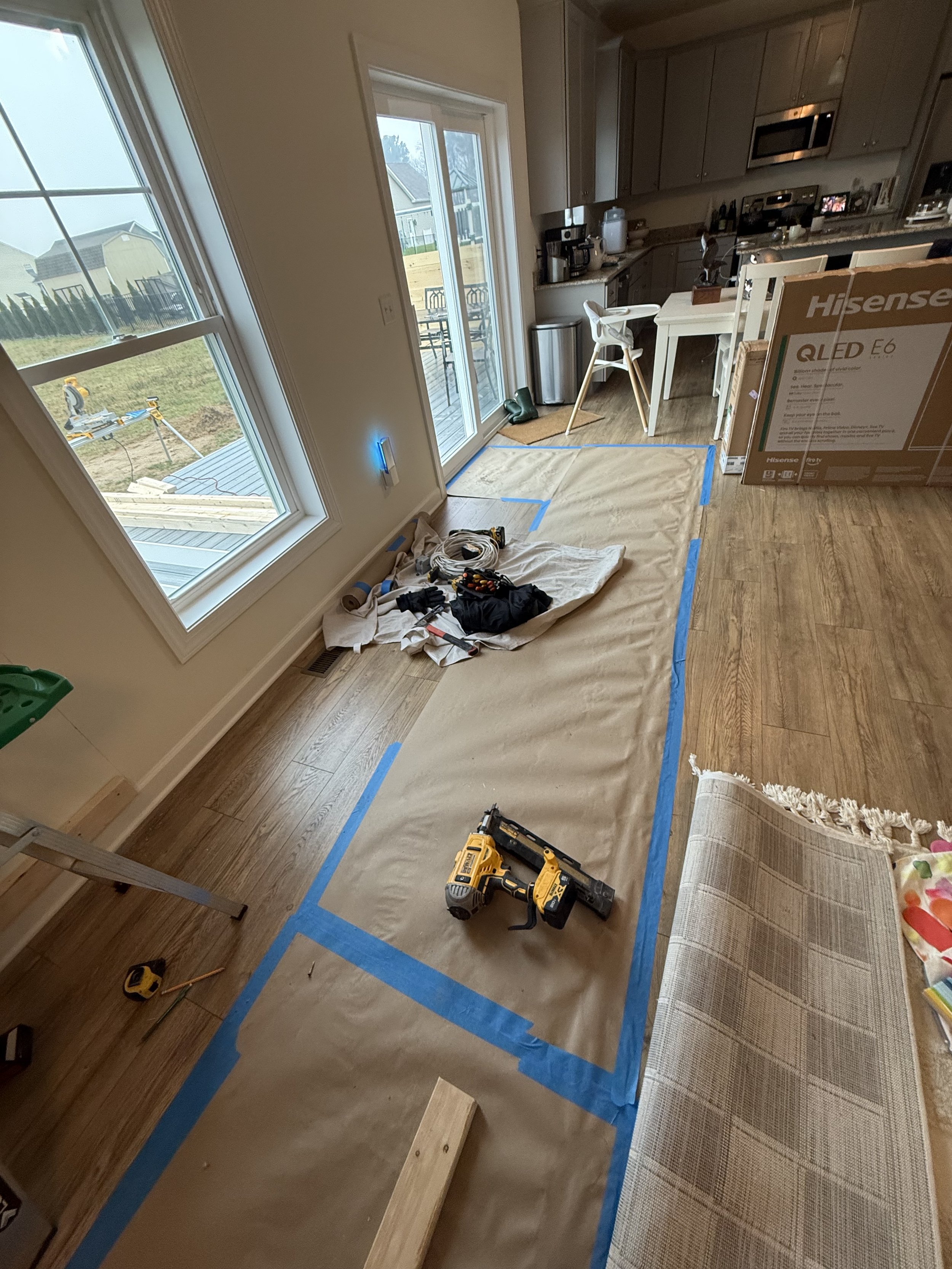 Living room under renovation with paper covering the floor, tools including a cordless drill, and a piece of wood. There are large windows, a sliding door leading to a deck, and kitchen cabinets in the background.