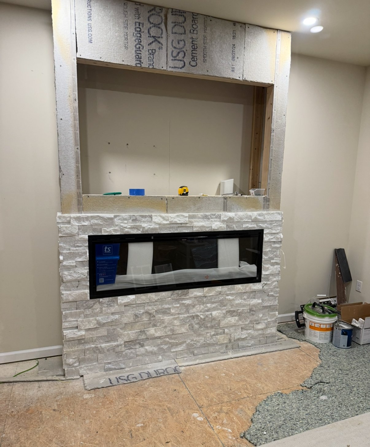 Ongoing interior construction of a wall with a fireplace, with partially finished stone veneer, tools, and construction materials visible.