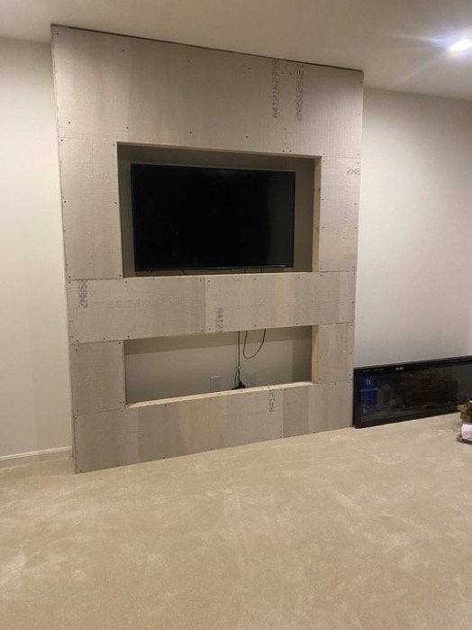 A partially built, custom wall with a large television opening and a smaller storage or display space below, in a room with unfinished construction.