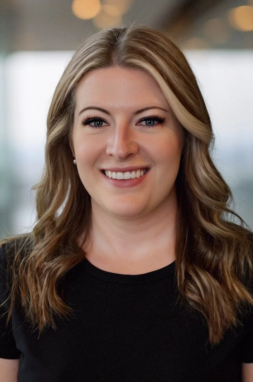 A woman with wavy honey-blonde hair, blue eyes, and a friendly smile, wearing a black shirt, in an indoor setting with blurred background lights.