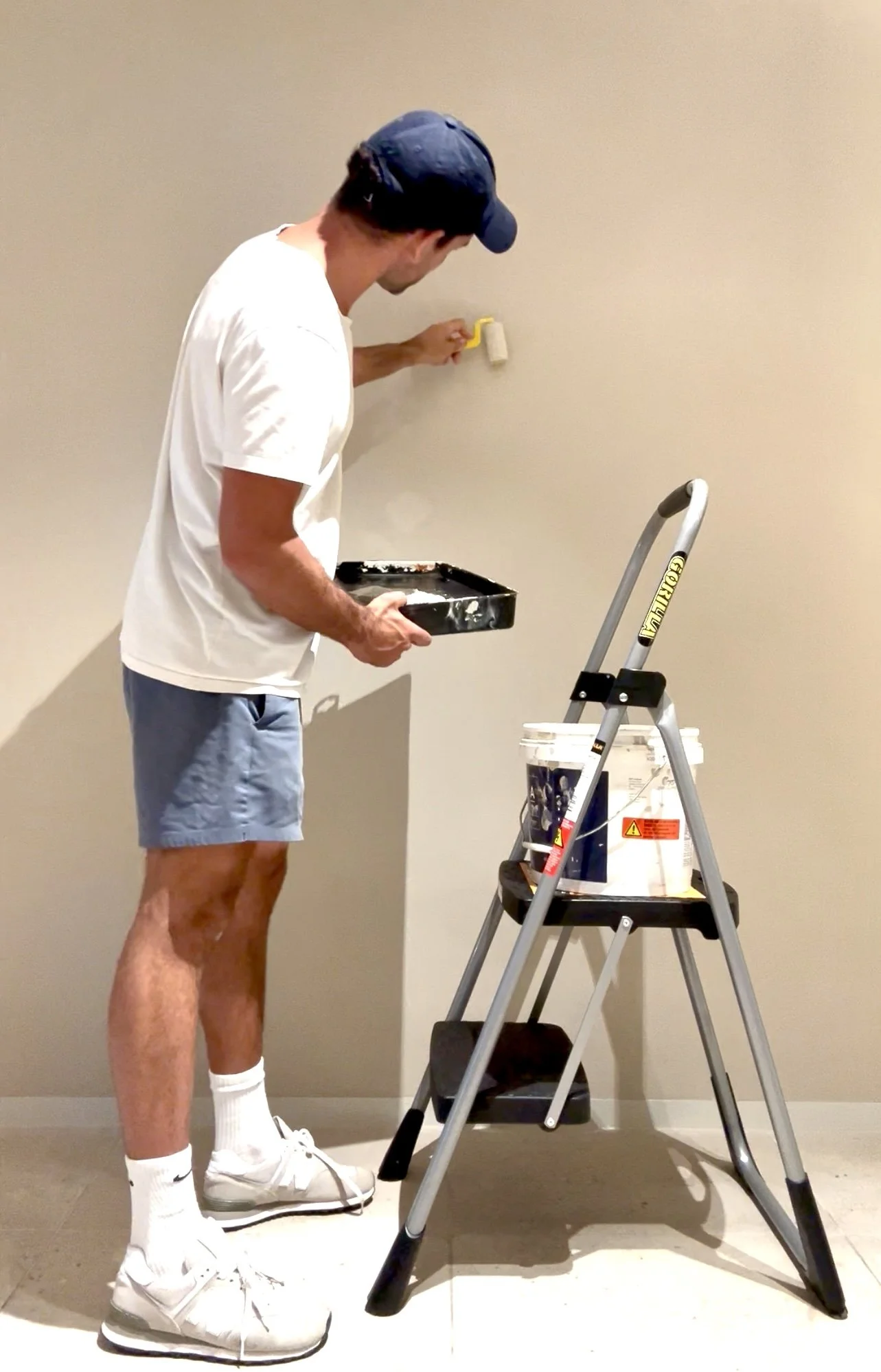 A man painting a wall with a paint roller, standing on a stepladder with a tray of paint in hand, dressed casually in shorts, a white t-shirt, white sneakers, white socks, and a baseball cap.