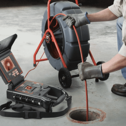 A person using a sewer camera inspection device with a red cable going into a floor drain. The setup includes a monitor displaying the camera feed and a cable reel. The person is wearing gloves and utilitarian clothing.