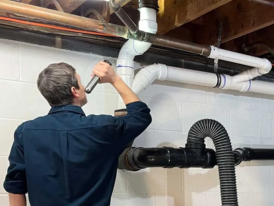 Person inspecting plumbing pipes in a basement with a flashlight.