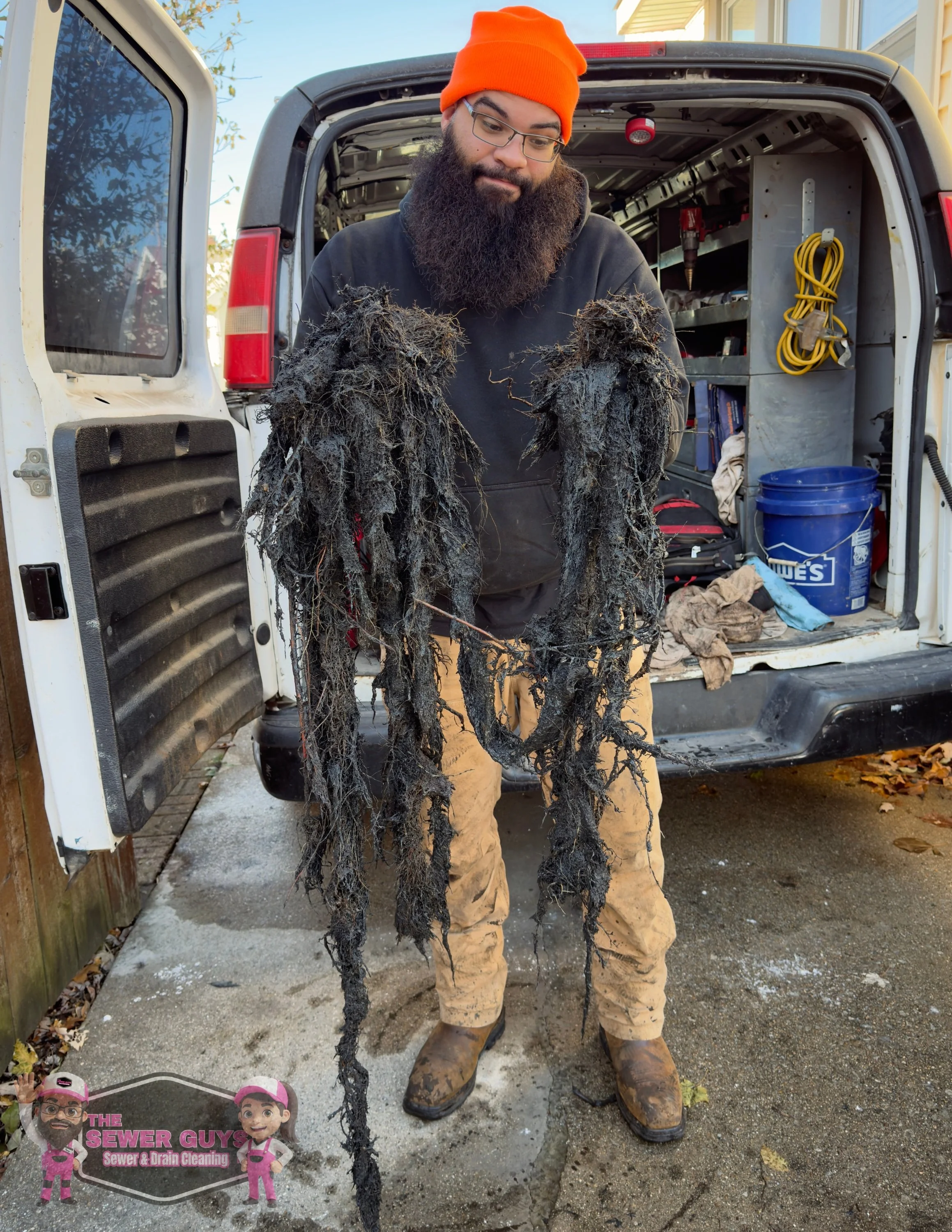 Man in black hoodie, tan pants, and orange beanie holding matted, dirty, and tangled root debris from a drain cleaning job, standing in front of a van with tools and equipment for sewer cleaning, with a logo that reads 'The Sewer Guys' in the bottom left corner.