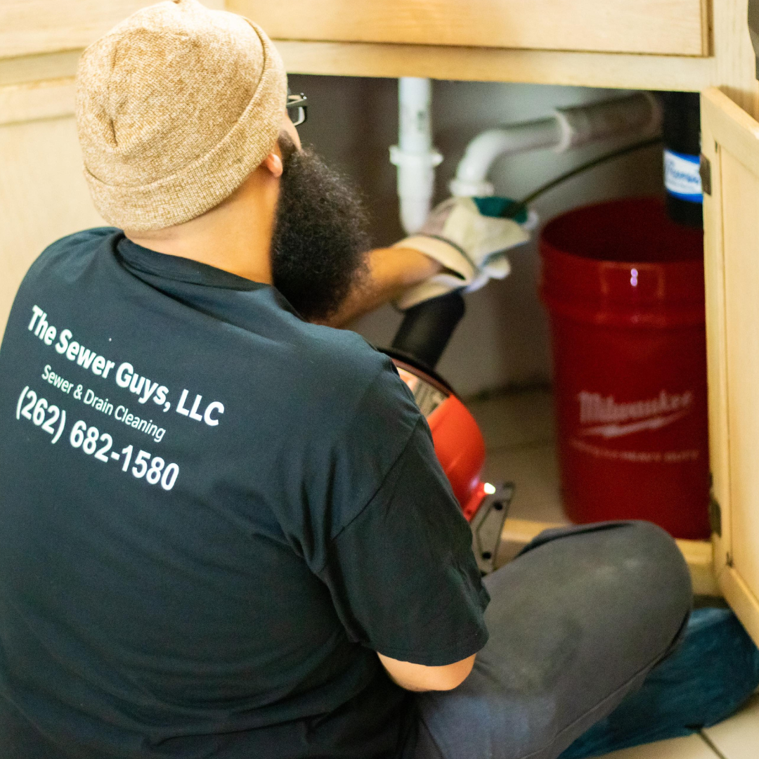 A person wearing a brown beanie and a dark t-shirt with company contact information, working under a kitchen sink with plumbing tools and a red bucket nearby.