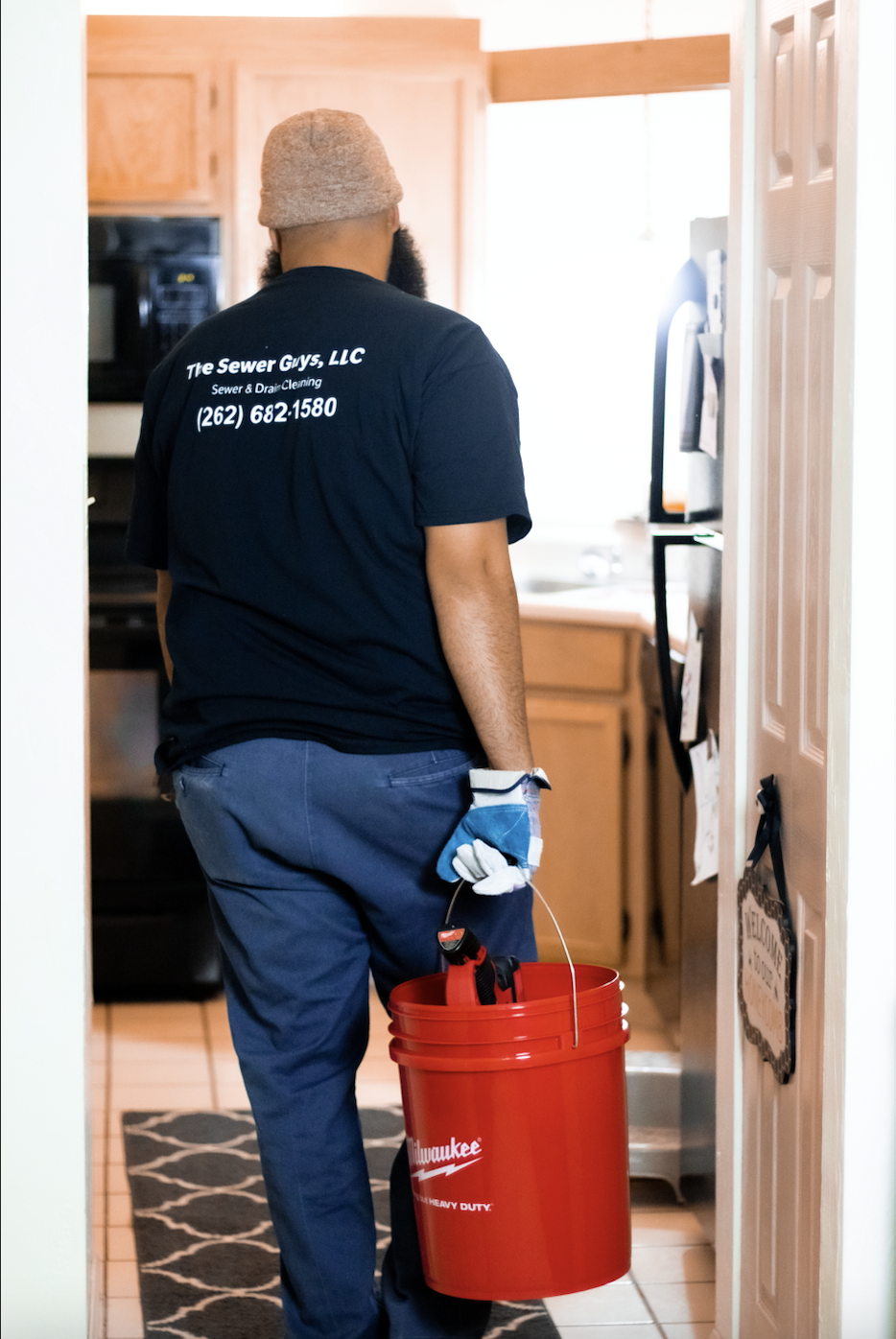A plumber standing in a kitchen, holding a red bucket with tools, wearing a dark t-shirt with company information and phone number on the back, a beige cap, and blue pants.