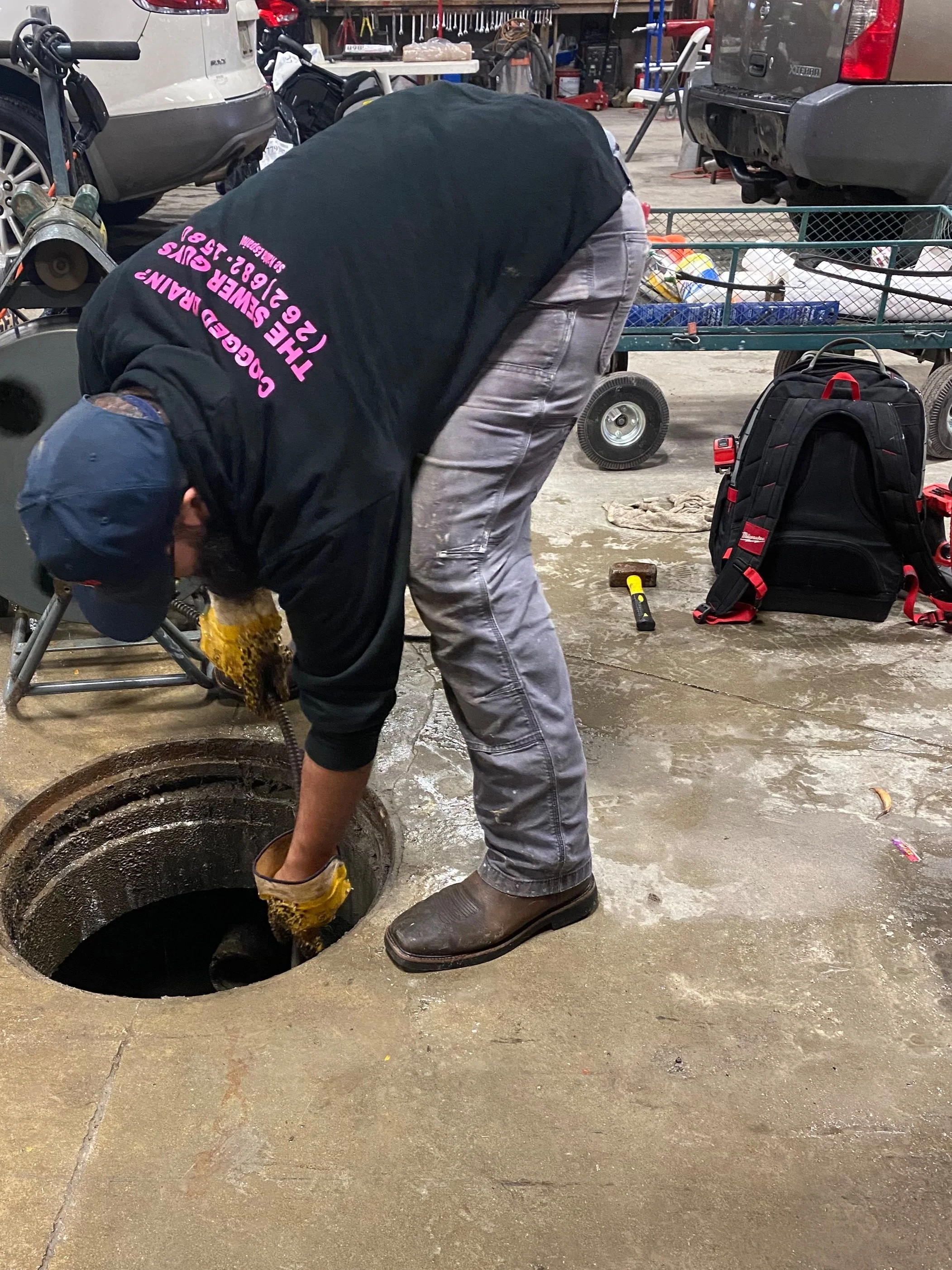 Person working on a drain or manhole in a garage, wearing gloves and boots, with tools and equipment around.