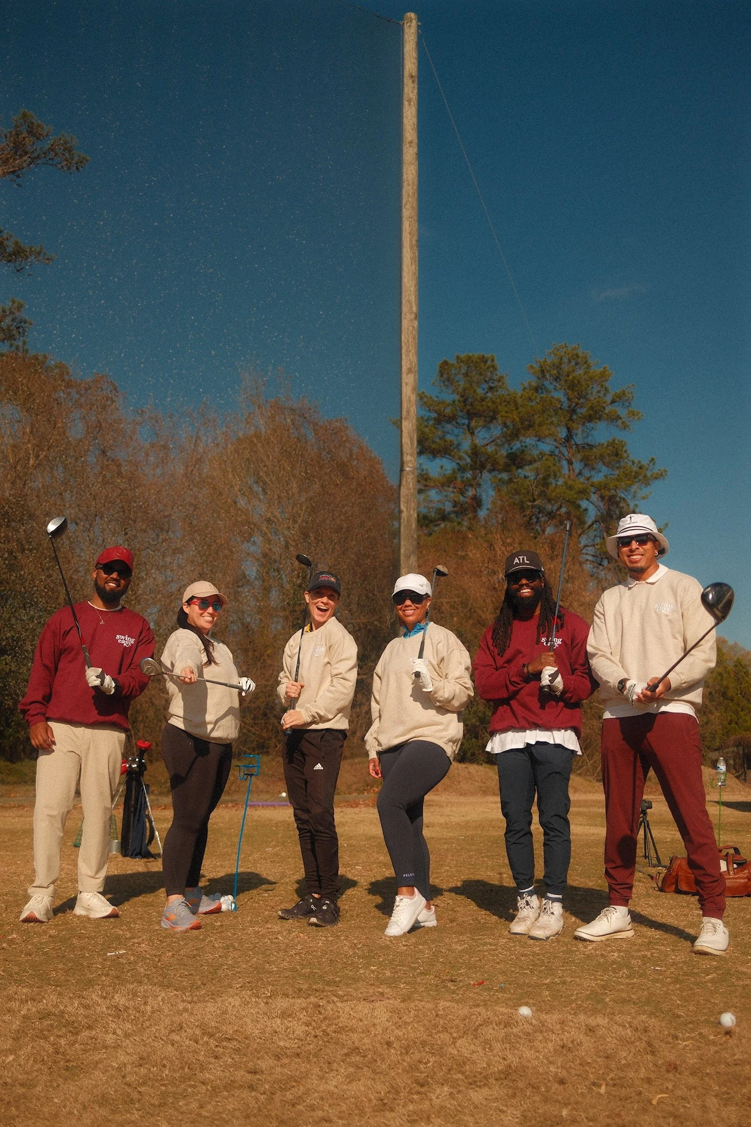 A diverse group of six friends standing on a golf course, holding clubs and smiling at the camera, with a clear blue sky and trees in the background, participating in a swing easy. golf clinic for beginners and high-handicap golfers.