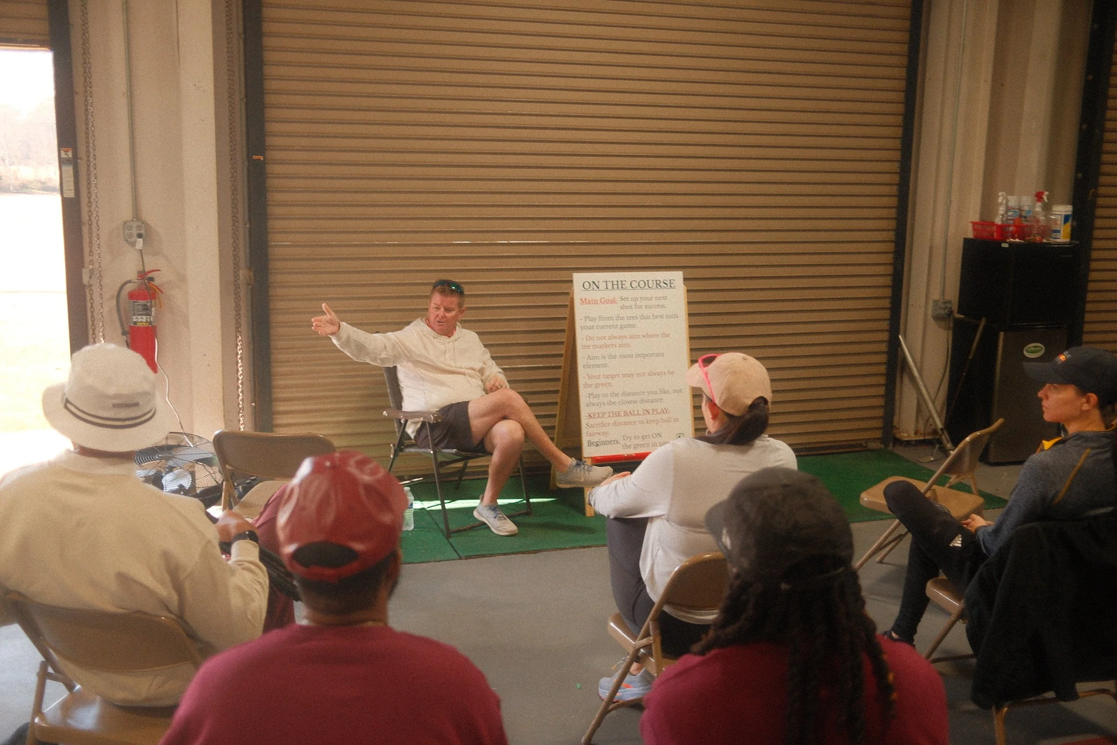 A group of people listening to a golf instructor in front of a large sign with instructions; the speaker is seated and gesturing while participants enjoy a swing easy. golf clinic for beginners and high-handicap golfers.