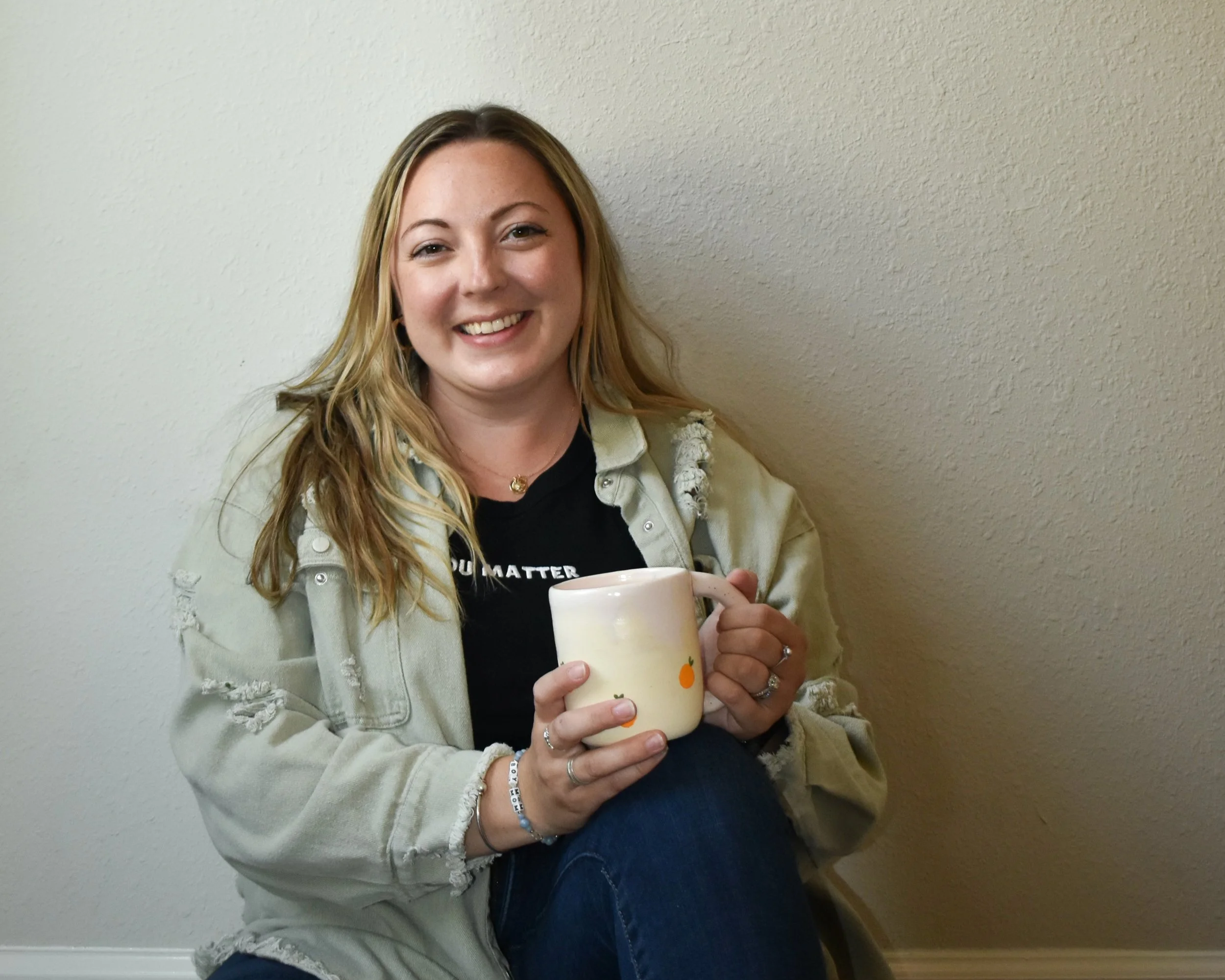 A smiling woman with blonde hair sitting against a beige wall, holding a white ceramic mug with an orange design, wearing a black shirt and a ripped beige jacket.