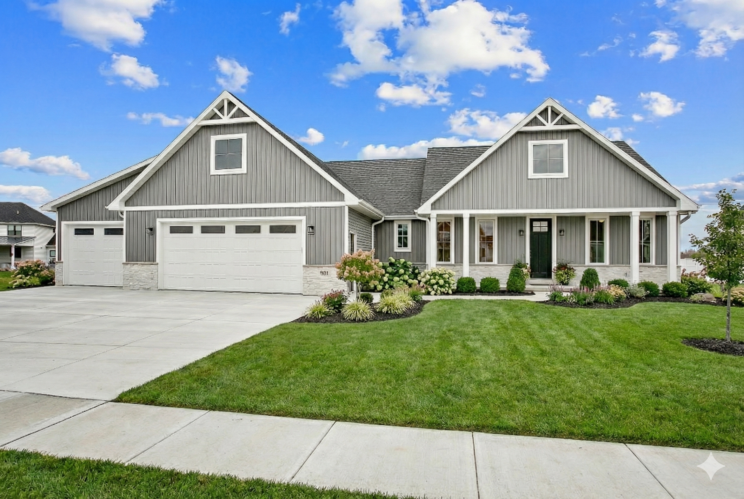 A modern gray house with a front porch, white trim, and a three-car garage, surrounded by a well-manicured lawn and landscaped garden, with a bright blue sky and some clouds overhead.