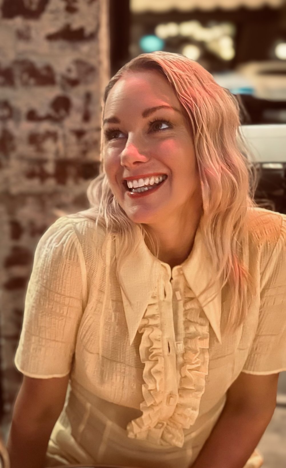 A headshot of Anita Fitzgerald smiling with blonde wavy hair sitting indoors near a brick wall.