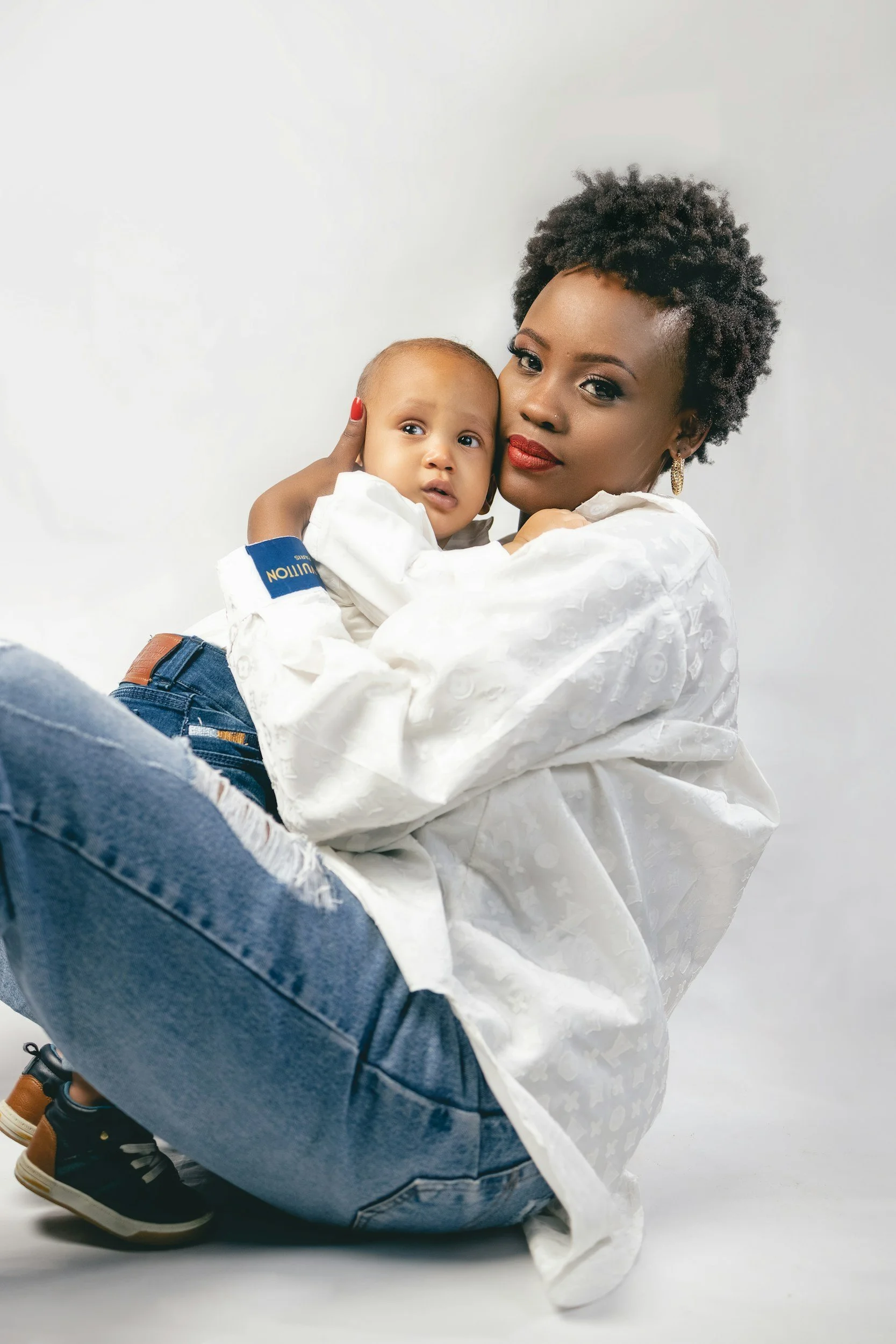 A woman with short curly hair and red lipstick holding a young child, both looking at the camera, against a plain white background.