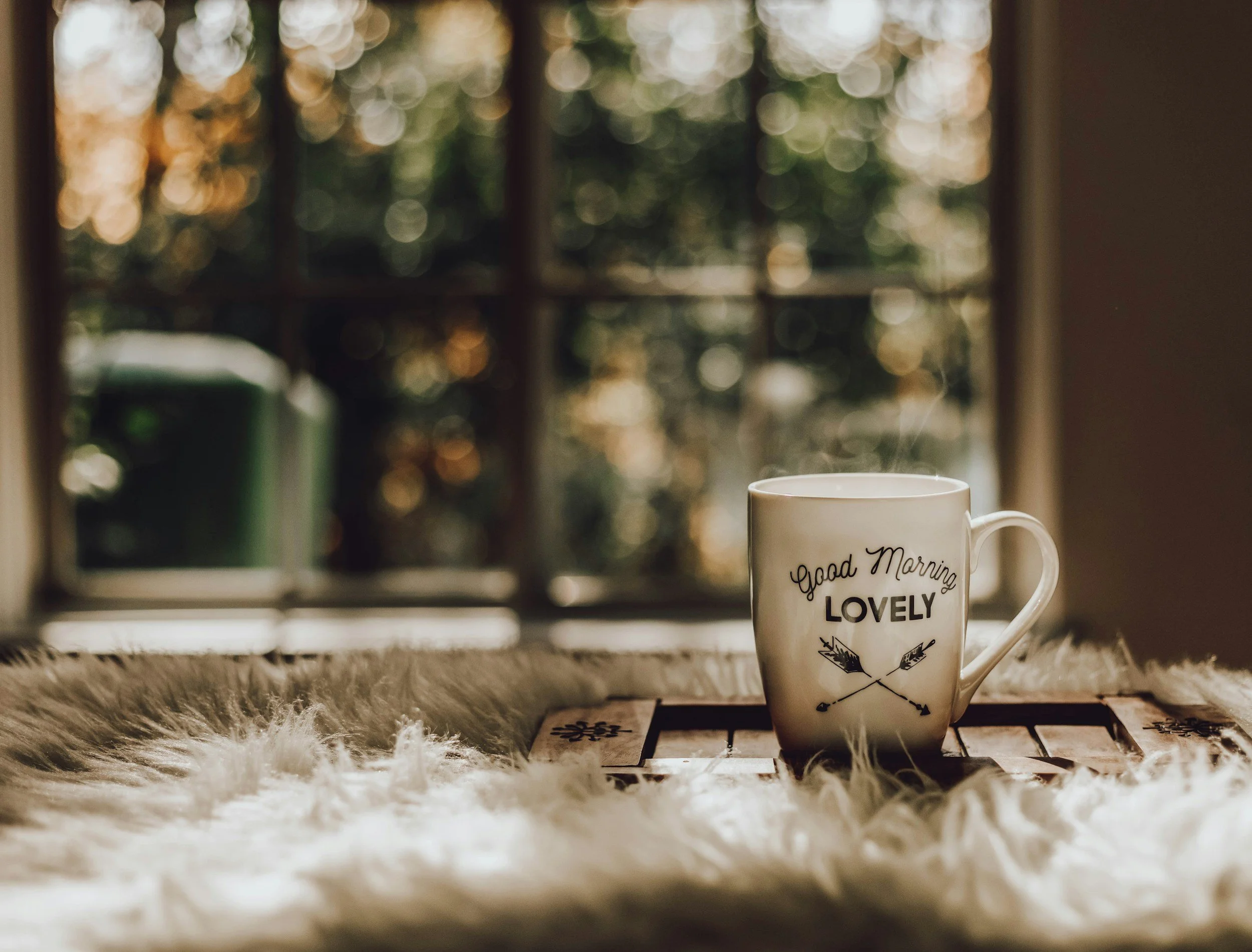 A white coffee mug with the words "Good Morning Lovely" and crossed arrows design, placed on a wooden surface with a fuzzy white rug, in front of a window with blurred autumn foliage outside, steam rising from the mug.