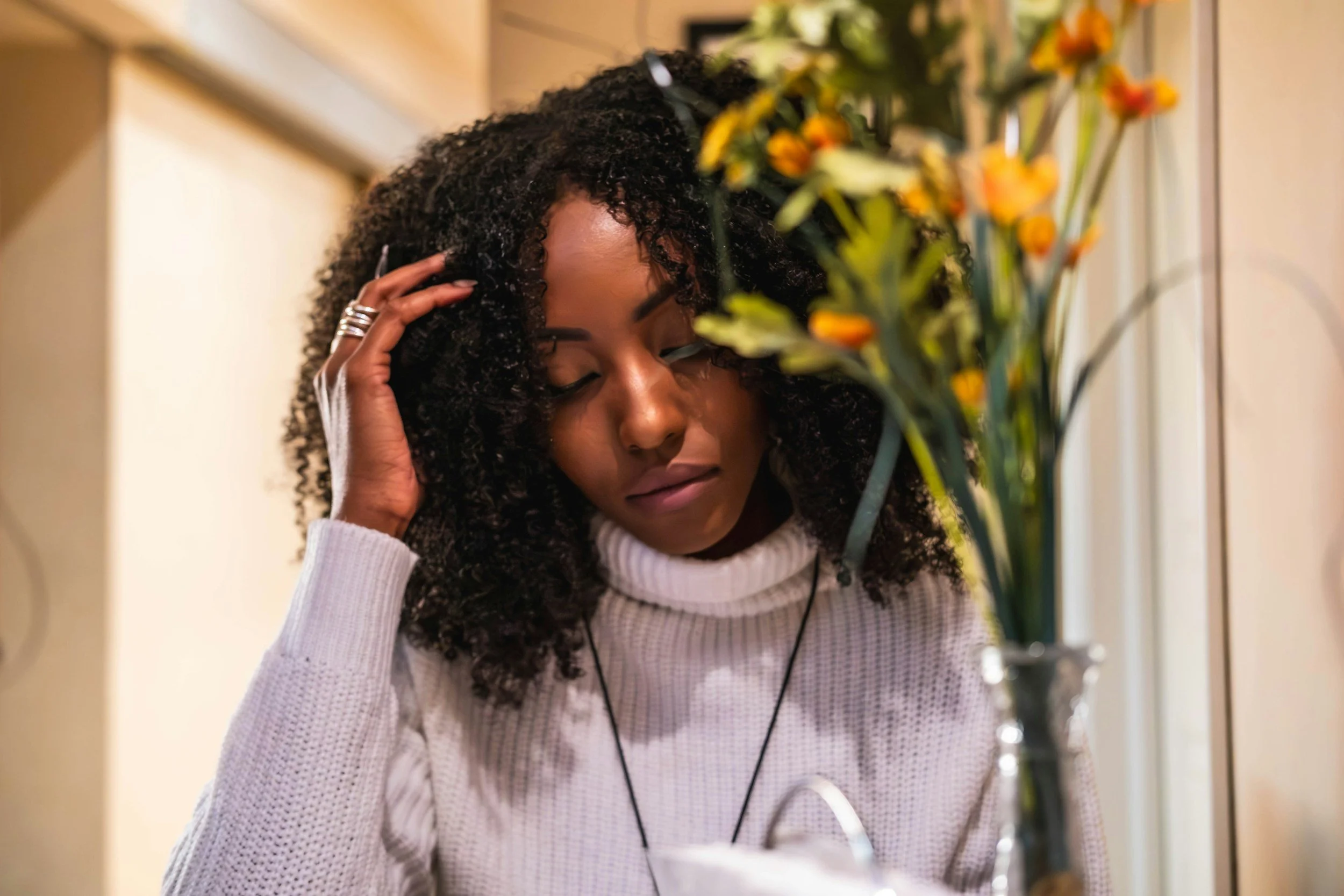 A woman with curly hair wearing a white sweater and listening to music stands near a vase of yellow and orange flowers, with her eyes closed and hand touching her hair.