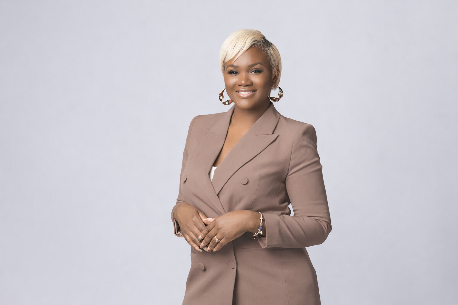 A woman with short, blonde hair wearing a beige blazer and hoop earrings, smiling and standing against a light gray background.