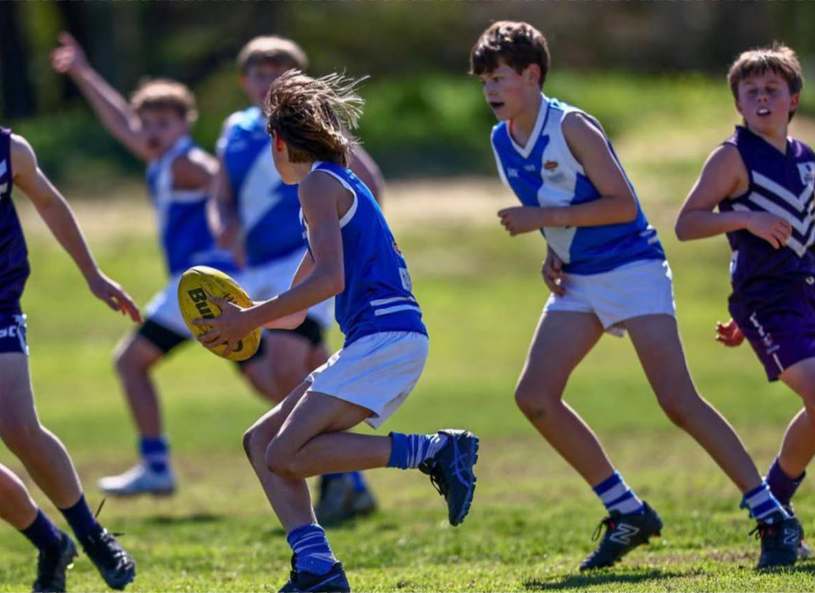 Children playing Australian rules football on a grassy field, with some running and one holding a yellow football.