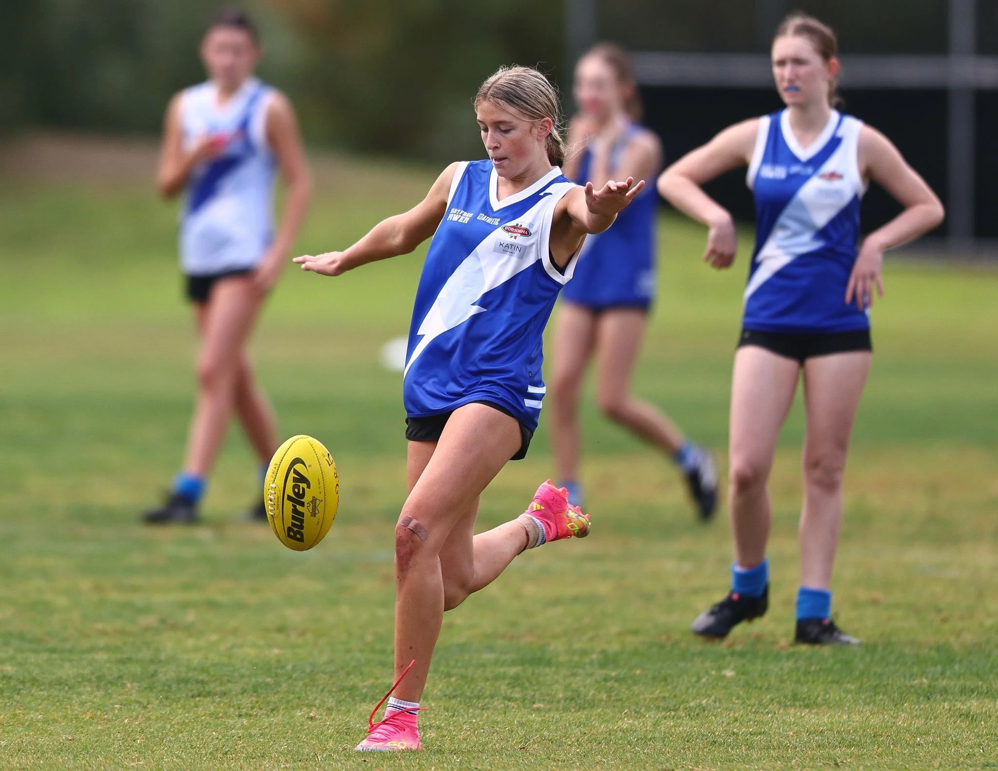 A girl in a blue sports uniform kicking a yellow football on a grassy field, with three other girls in similar uniforms watching in the background.