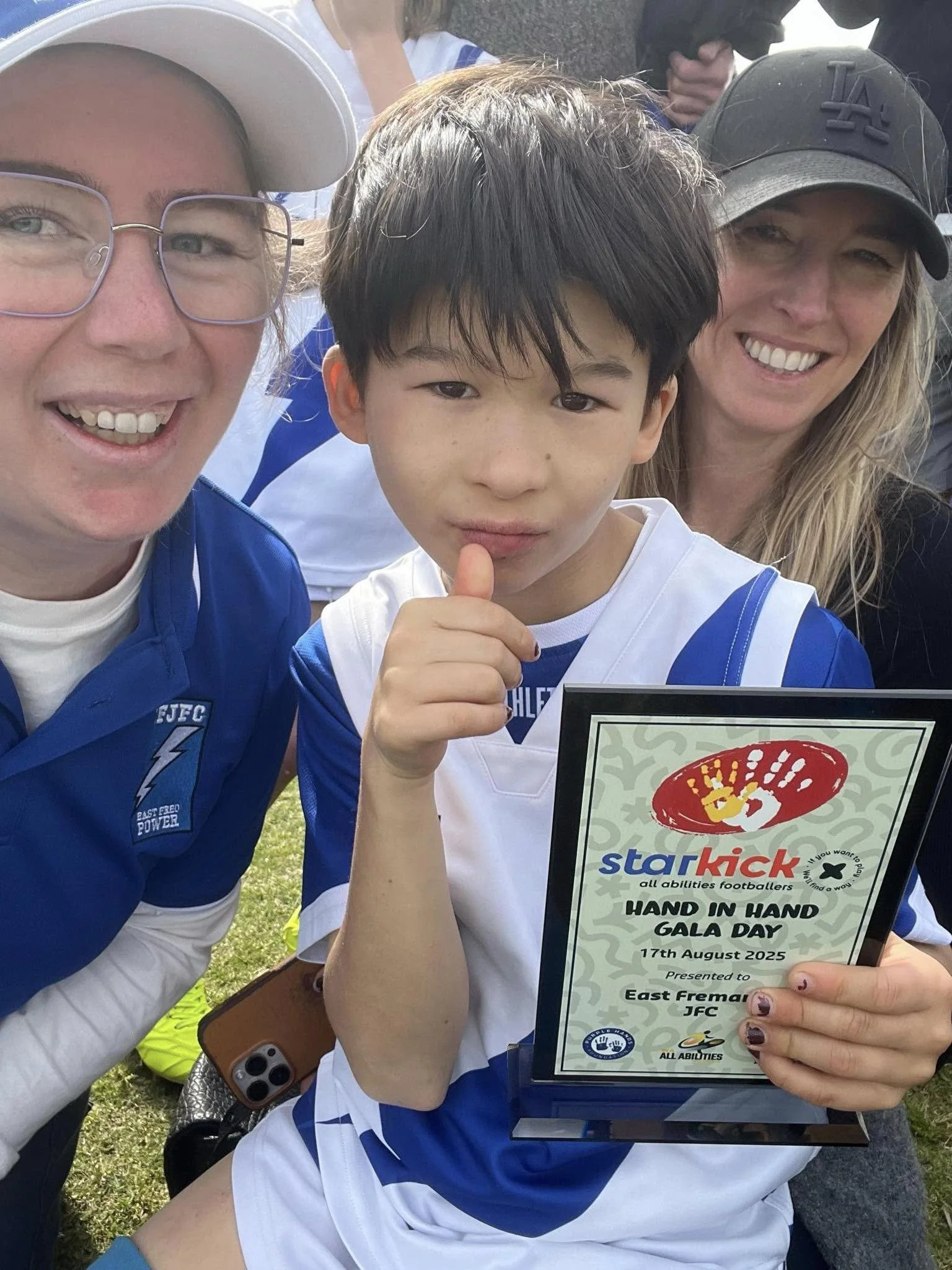 Young boy holding a plaque for Hand in Hand Gala Day at a football event, flanked by two smiling adults, outdoors on grass.
