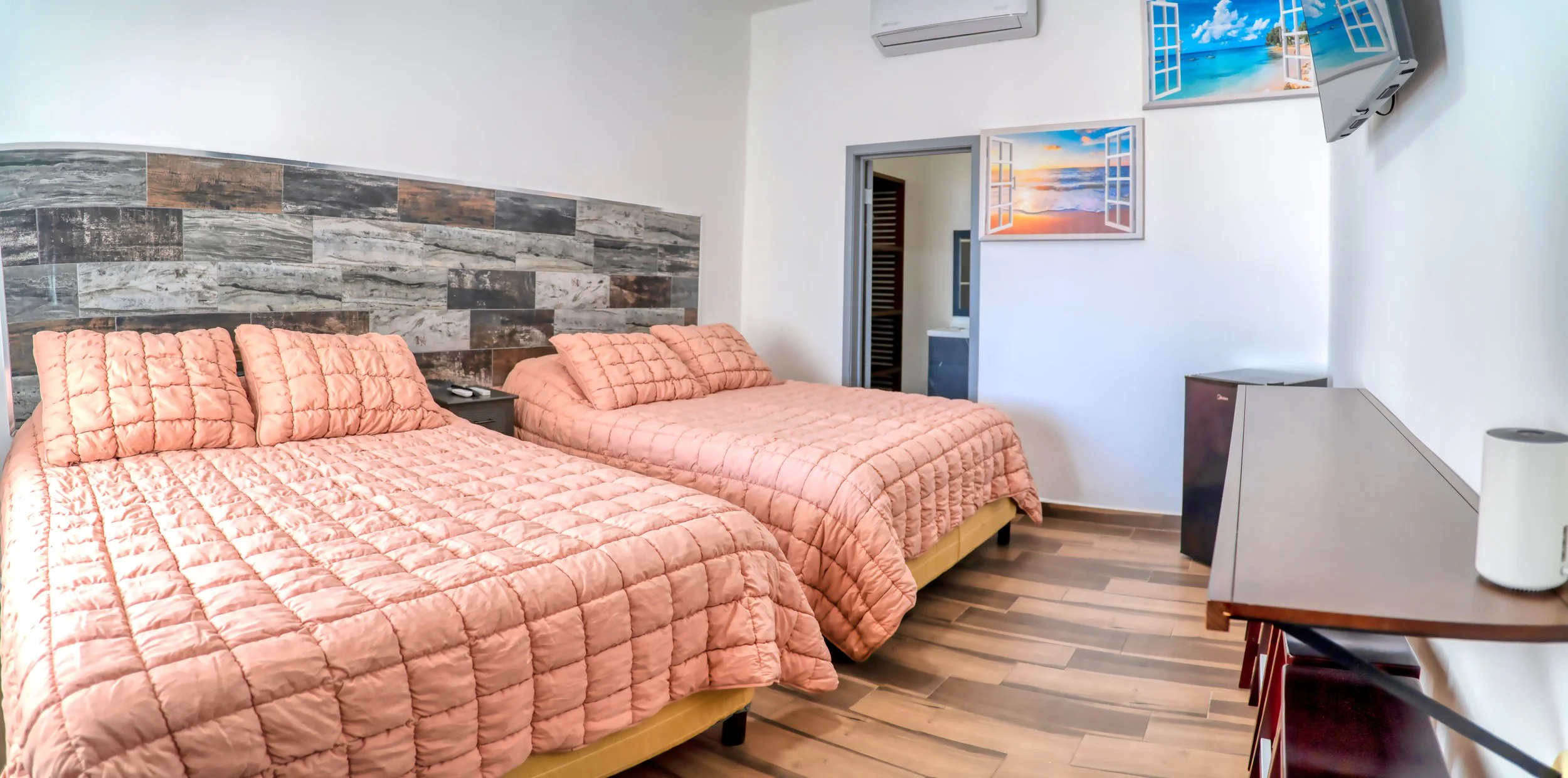 Habitación con dos camas individuales con colchas rosas, cabecera de madera con detalles de piedra, televisión en la pared, muebles y decoración moderna.