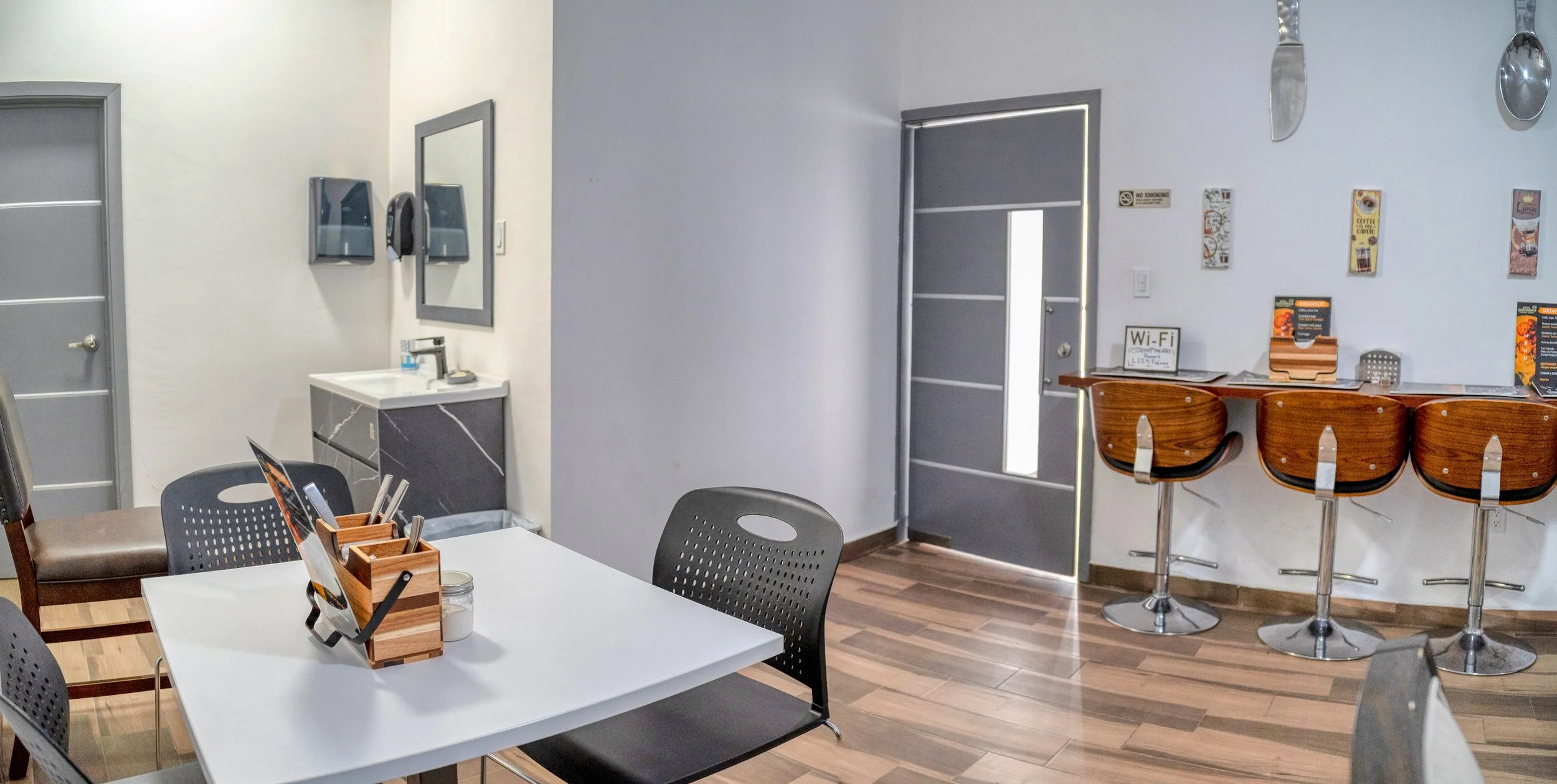 Interior de una cafetería con mesas y sillas, barra con banquetas de madera, paredes blancas, decoraciones y señal de Wi-Fi