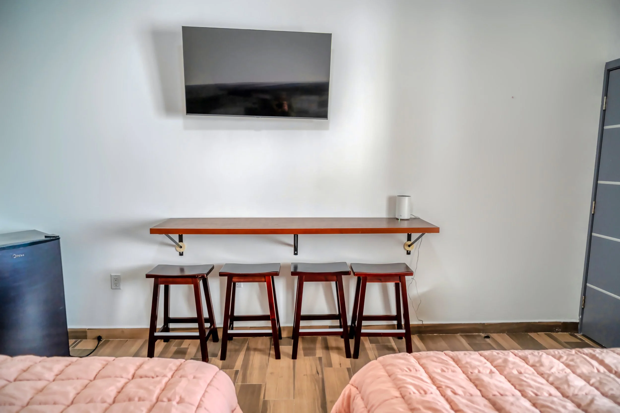Vista de un cuarto con dos camas con colchas rosadas, una mesa larga de madera con cuatro bancos, un televisor de pantalla plana en la pared, una mini nevera azul, una lámpara y una puerta gris en la esquina derecha.