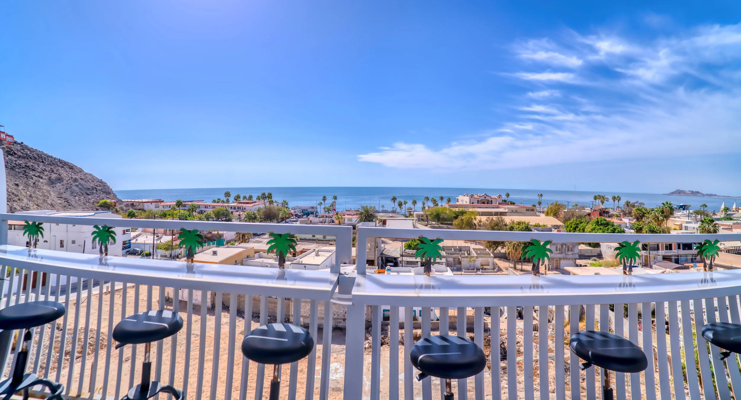 Vista desde un balcón con sillones y una barandilla decorada con palmeras, mirando hacia un paisaje de casas, palmeras, playa y el océano con cerros a lo lejos en un día soleado.