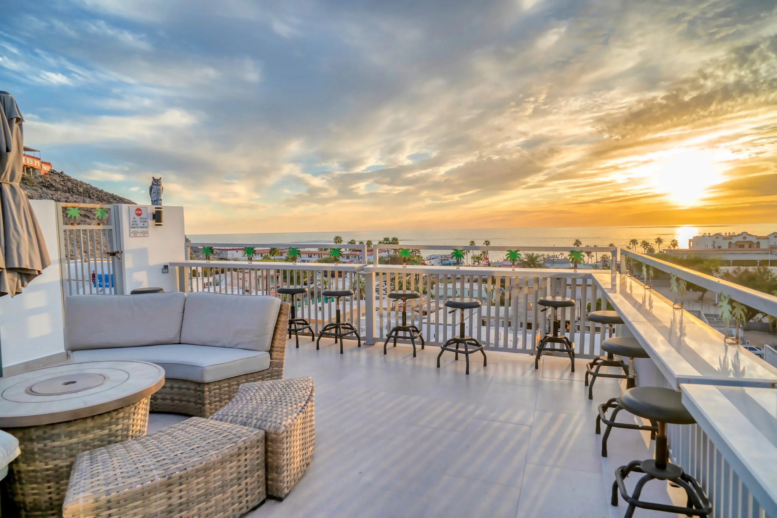Terraza al atardecer con vista al mar, sofá de mimbre, mesa redonda, sillas altas y decoración playera con palmeras en la distancia.