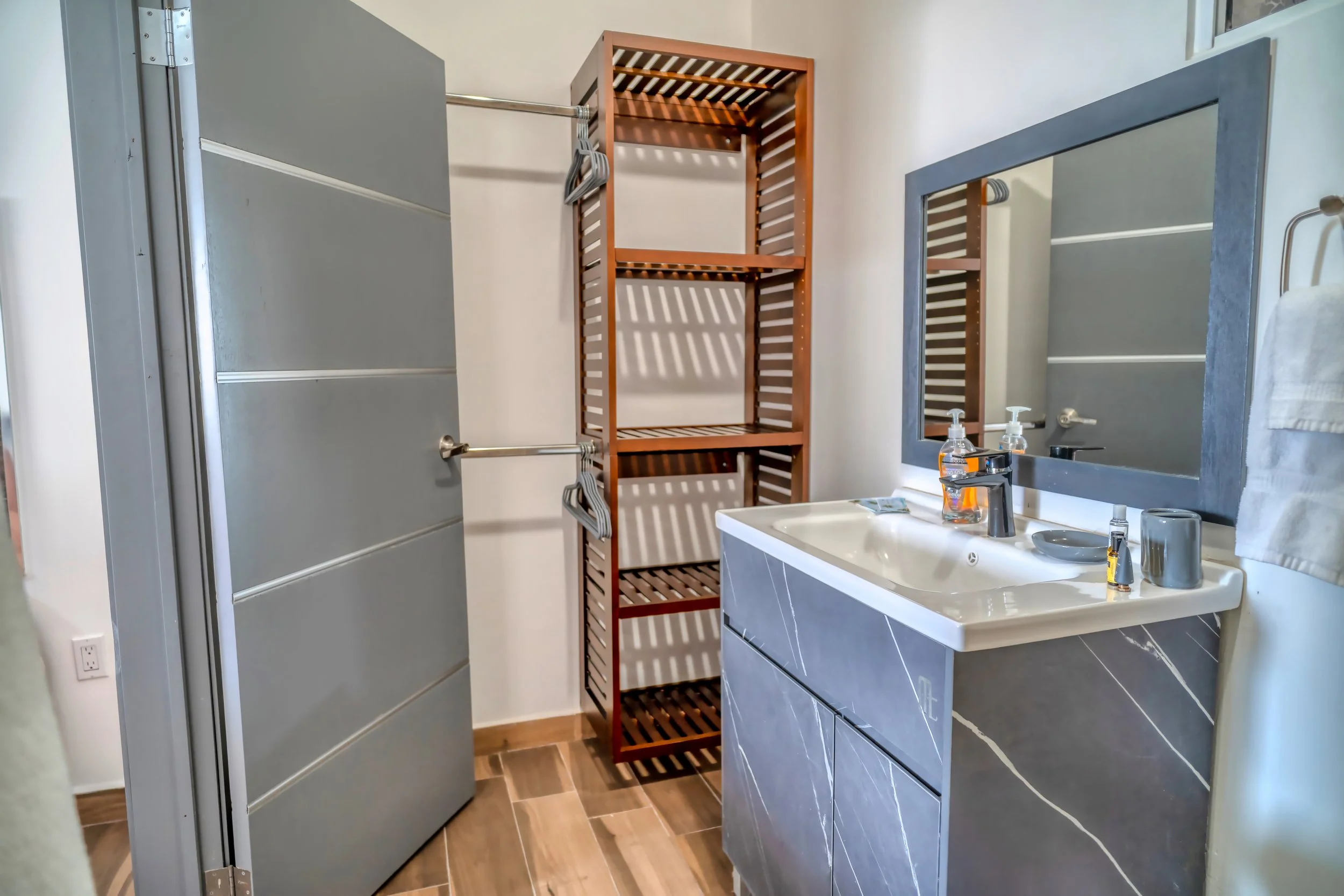 Un baño con espejo sobre lavabo, estantería de madera y ducha con puerta de metal.