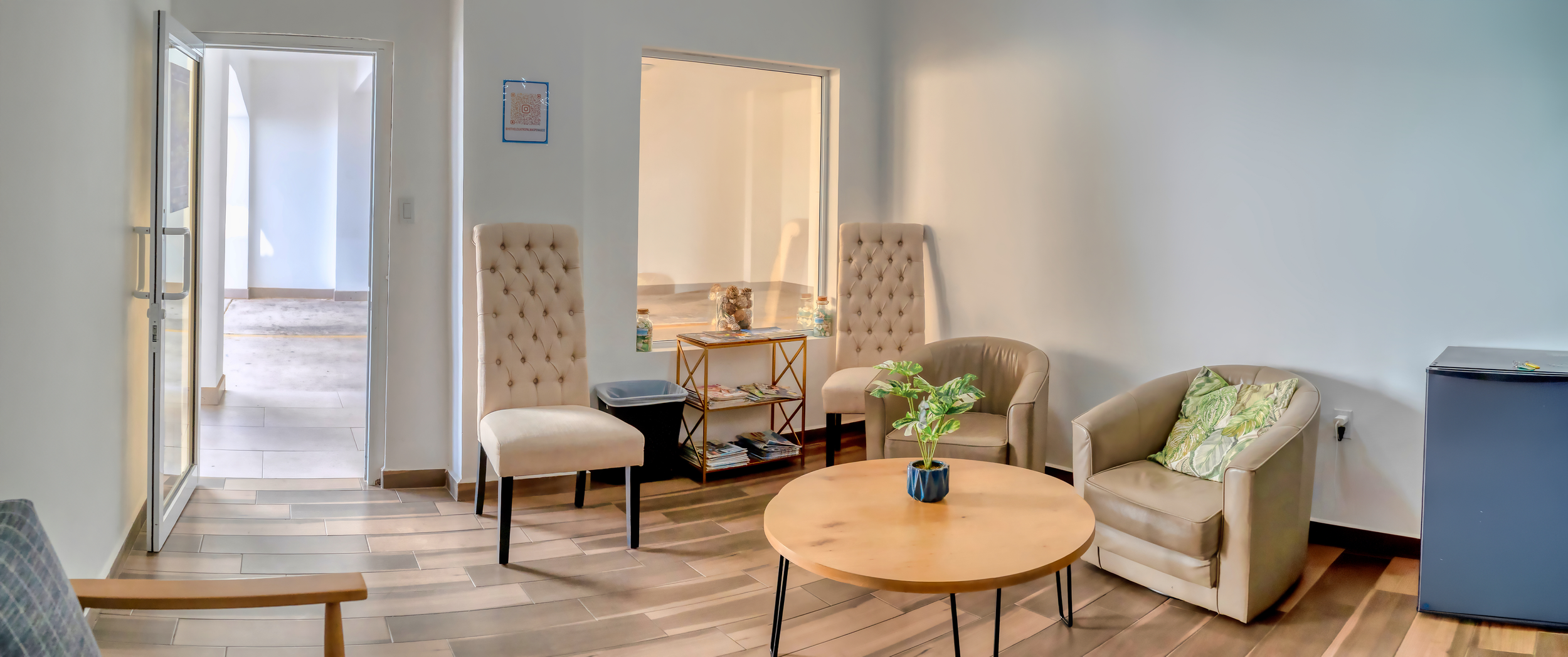 Sala de espera con sillones beige, mesa redonda de madera, plantas, y una pequeña estantería con revistas, en un ambiente doméstico o de oficina.