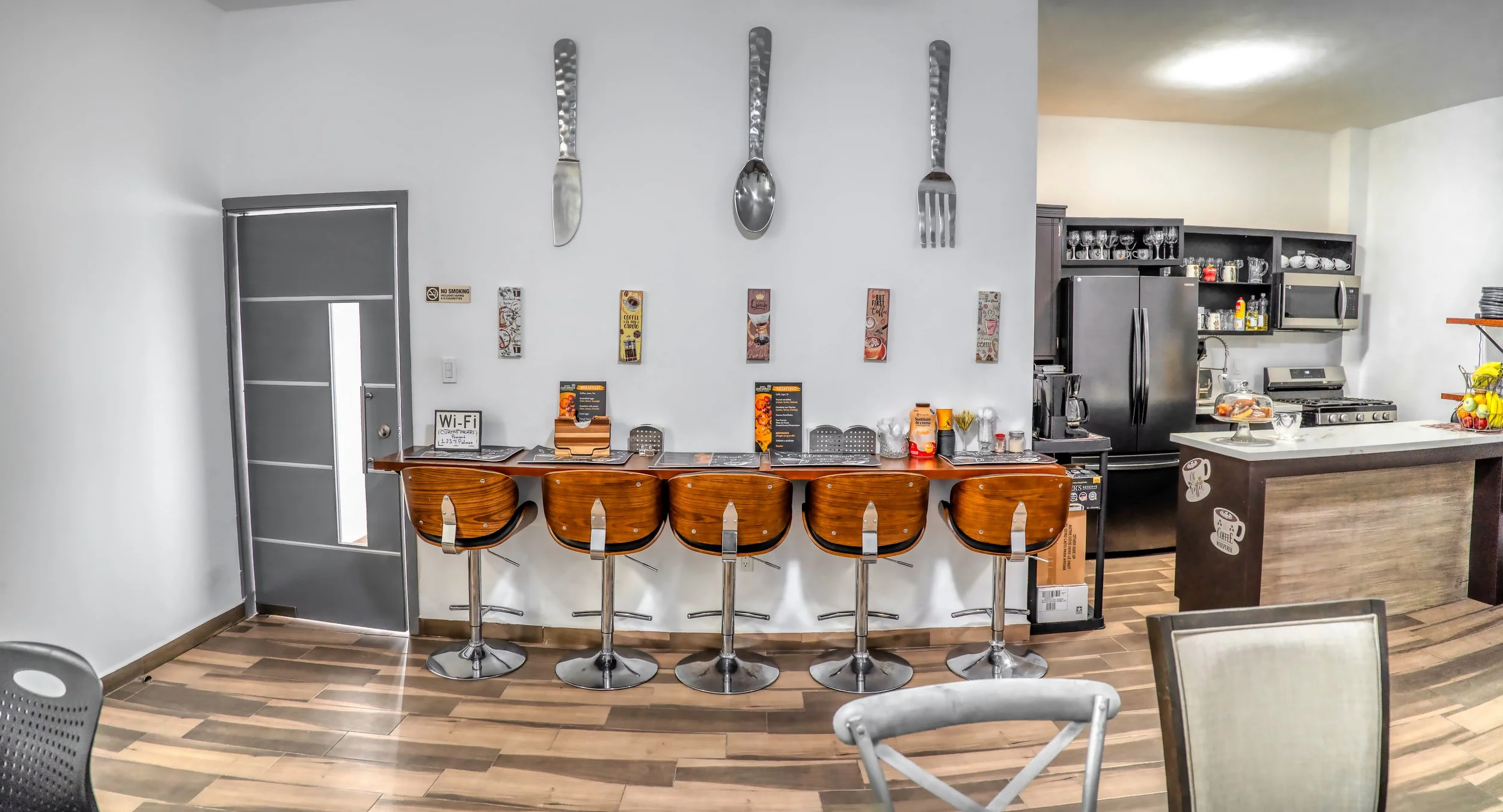Vista del interior de un café con una barra de madera con cinco sillas de bar de madera y metal. En la pared, utensilios grandes de cocina decorativos con forma de cuchillos, cucharas y tenedores. En el fondo, una cocina con electrodomésticos negros,
