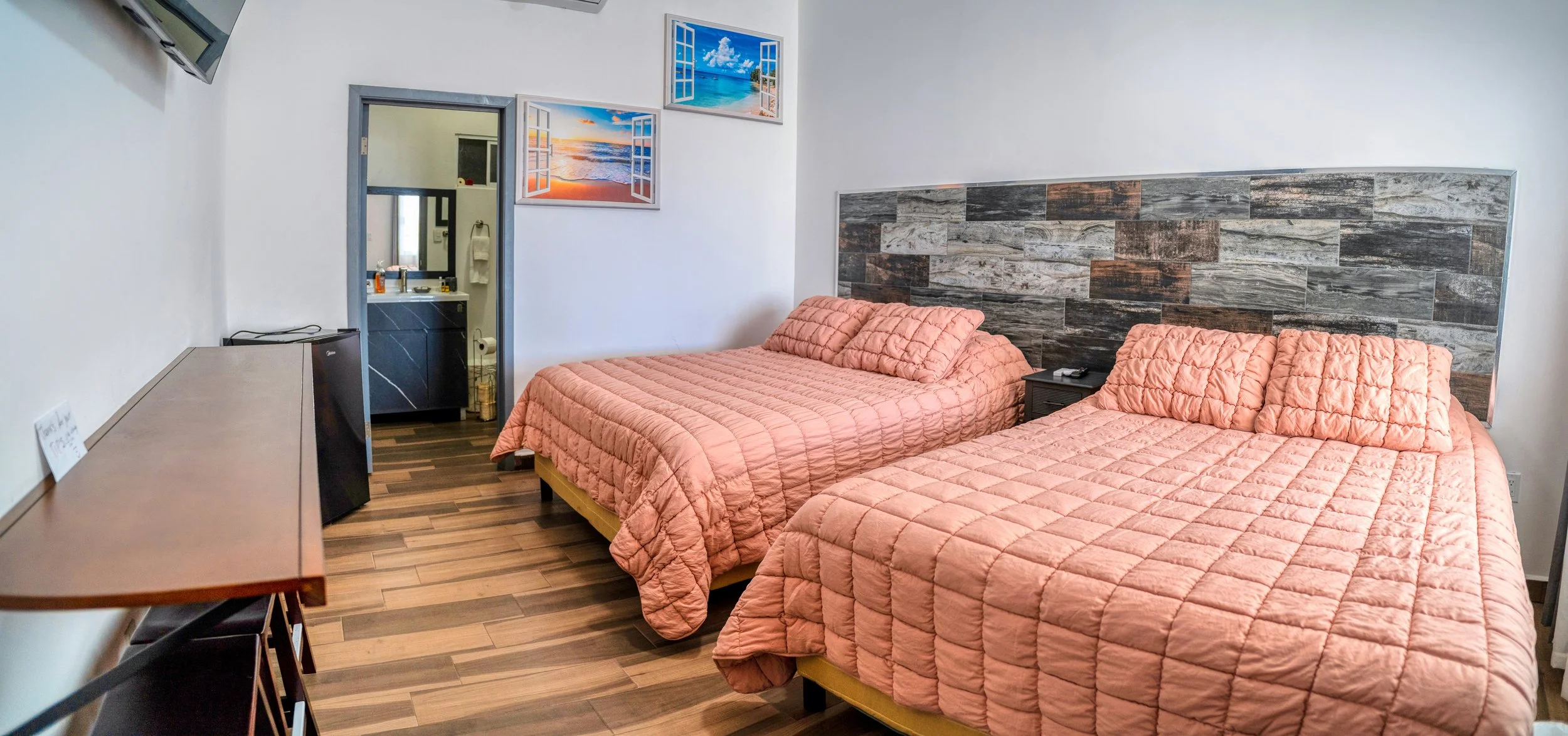 Habitación con dos camas con colchas rosas, cabecera de madera de tonos oscuros, una mesa de noche y una pequeña nevera. Decoración con cuadros de paisajes marinos y de atardeceres con ventanas abiertas. Tiene un cuarto de baño visible al fondo.