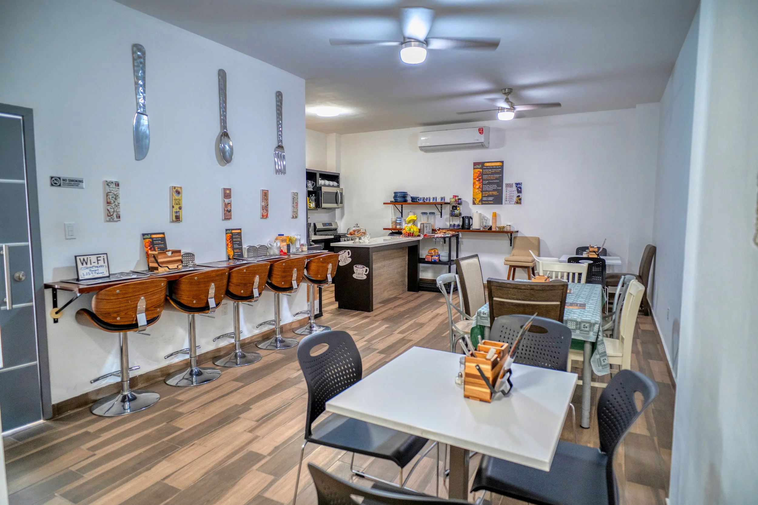 Sala de restaurante con mesas y sillas, área de servicio con barra, utensilios de comida en la pared, y una sección con máquinas y artículos de cocina al fondo.
