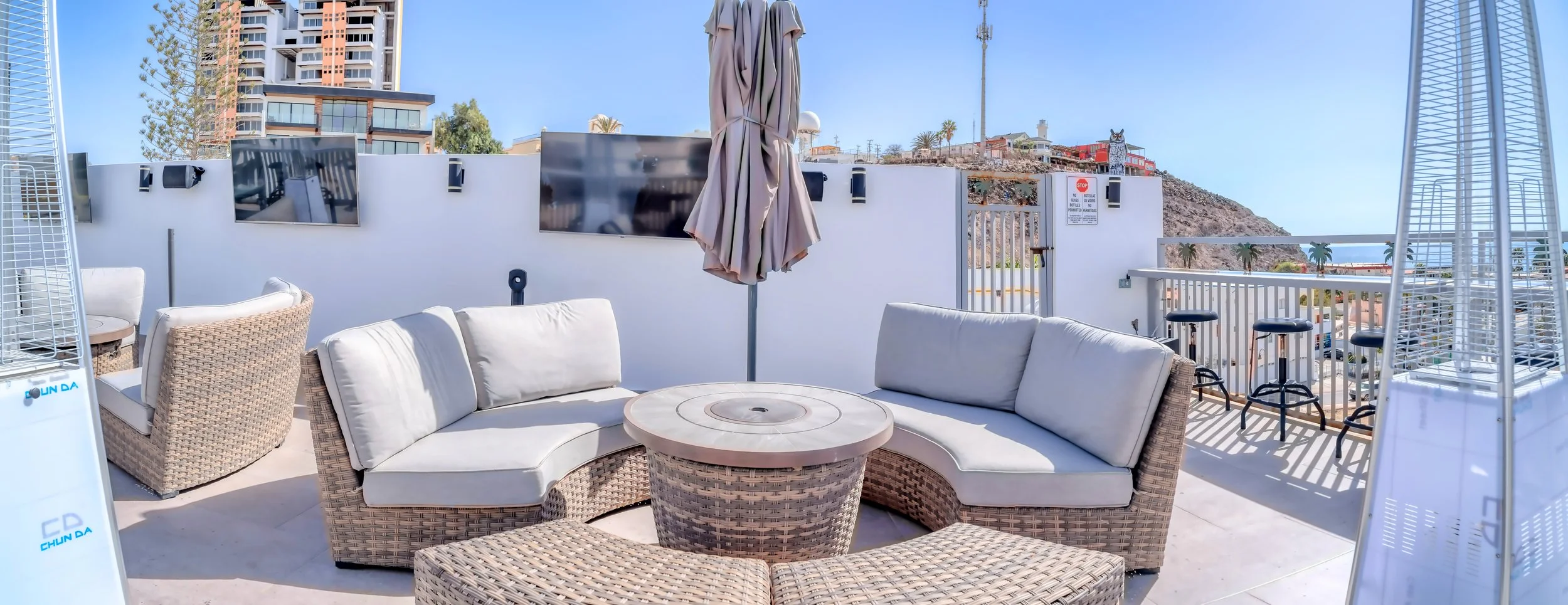 Terraza al aire libre con sillones blancos de mimbre y cojines en forma de L, mesa redonda de mimbre, sombrilla cerrada, toldos calefactores y vista a edificios y montañas en un día soleado.