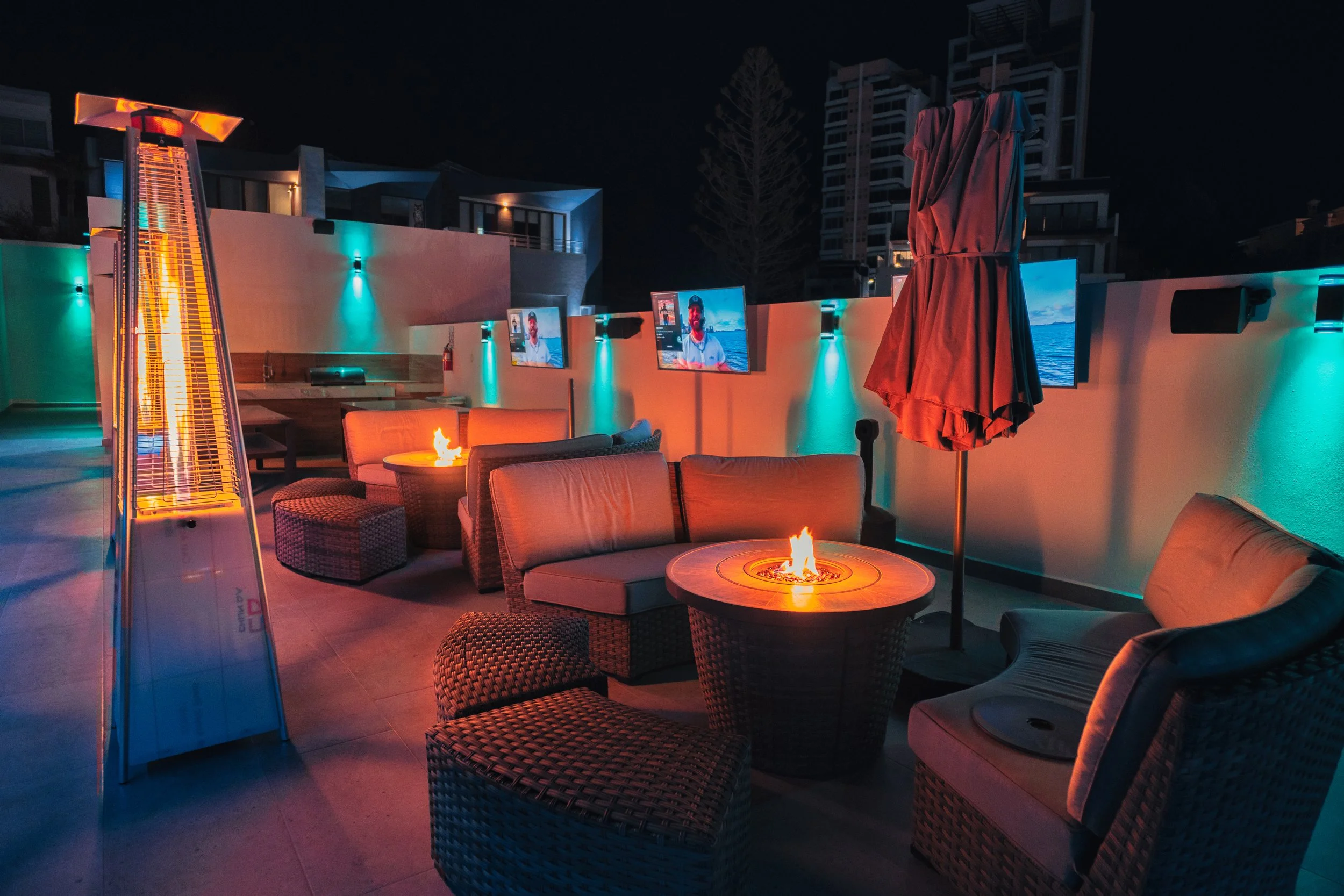 Terraza en la noche con muebles de mimbre, mesas con fuego, toldo cerrado, un calentador de exterior y pantallas de televisión, decorada con luces de colores teal y naranja.
