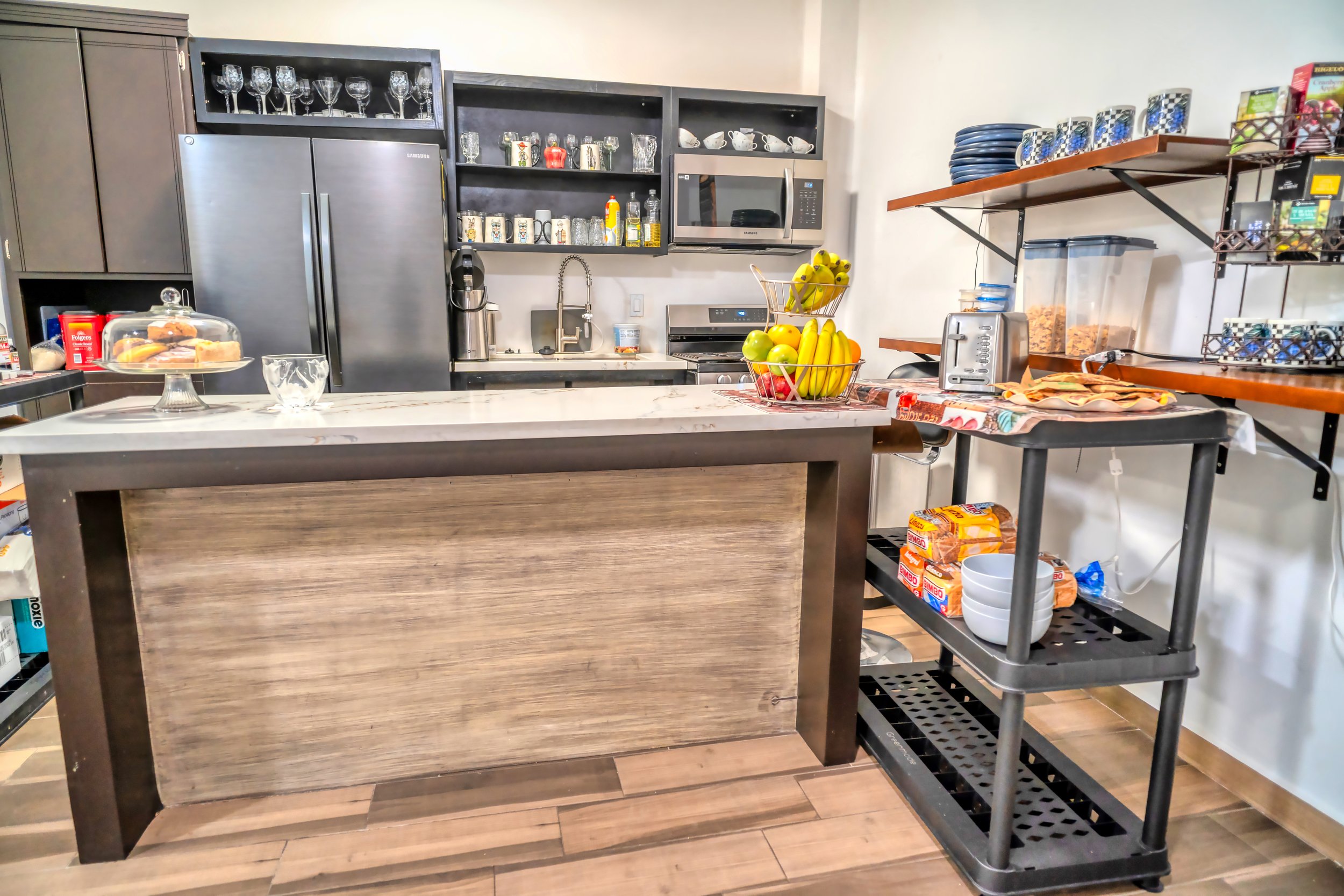 Cocina moderna con nevera de acero inoxidable, estantes con vasos, tazas y platos, una isla con fruta y un carrito de cocina con utensilios y comestibles, y una pared con repisas de madera con artículos de cocina y alimentos.